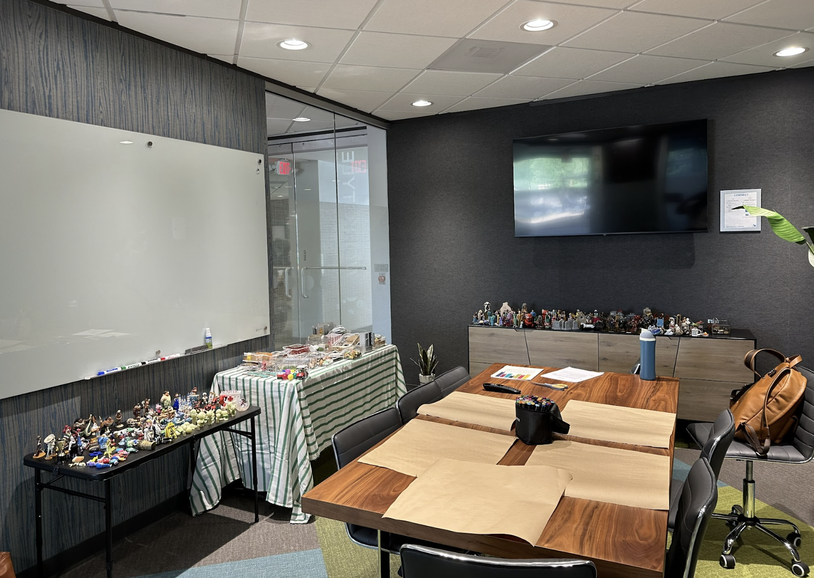 A small meeting room with a wooden table, several chairs, and a whiteboard. Walls are dark, with a TV mounted. Tables display assorted small items.