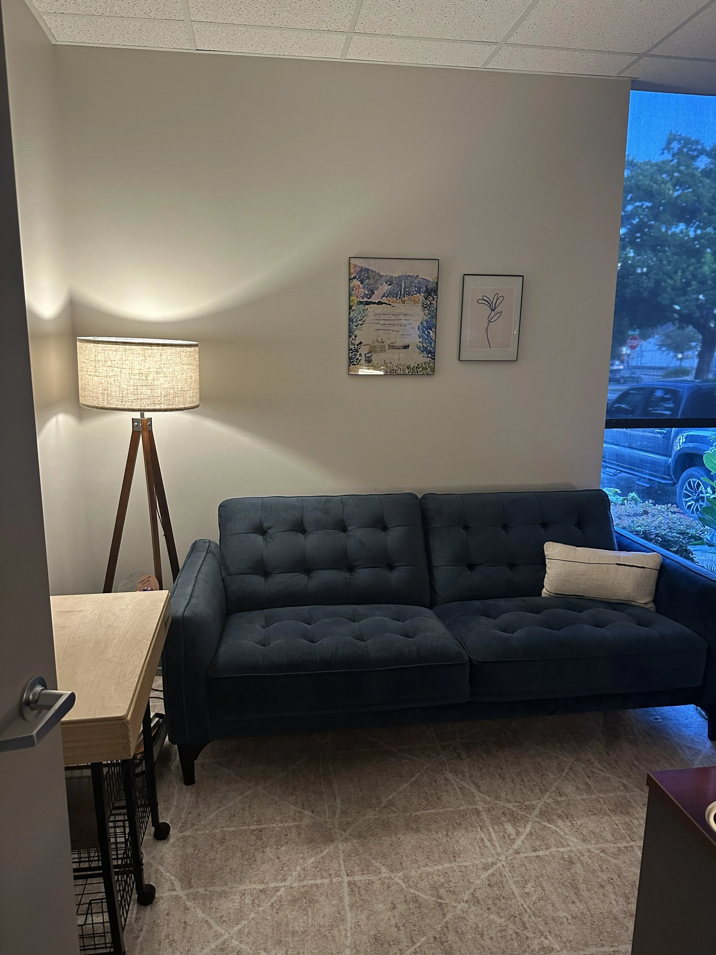 A modern decorated room with a blue sofa, a floor lamp, framed artwork on the wall, and a window showing trees outside.