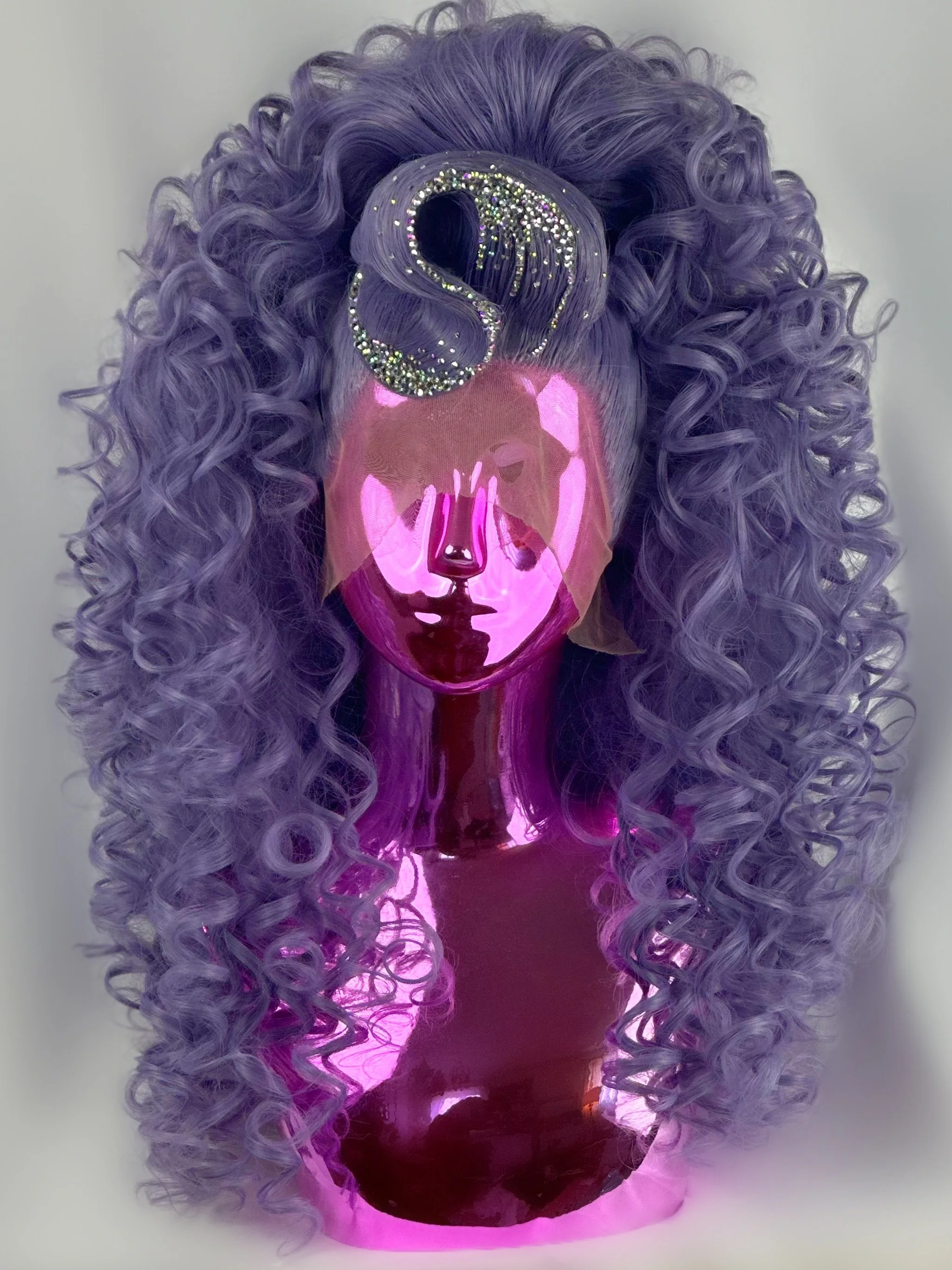 A mirror with a reflective, holographic pink surface, reflecting a person with curly purple hair styled in large ringlets, adorned with a rhinestone S-shaped hair clip.