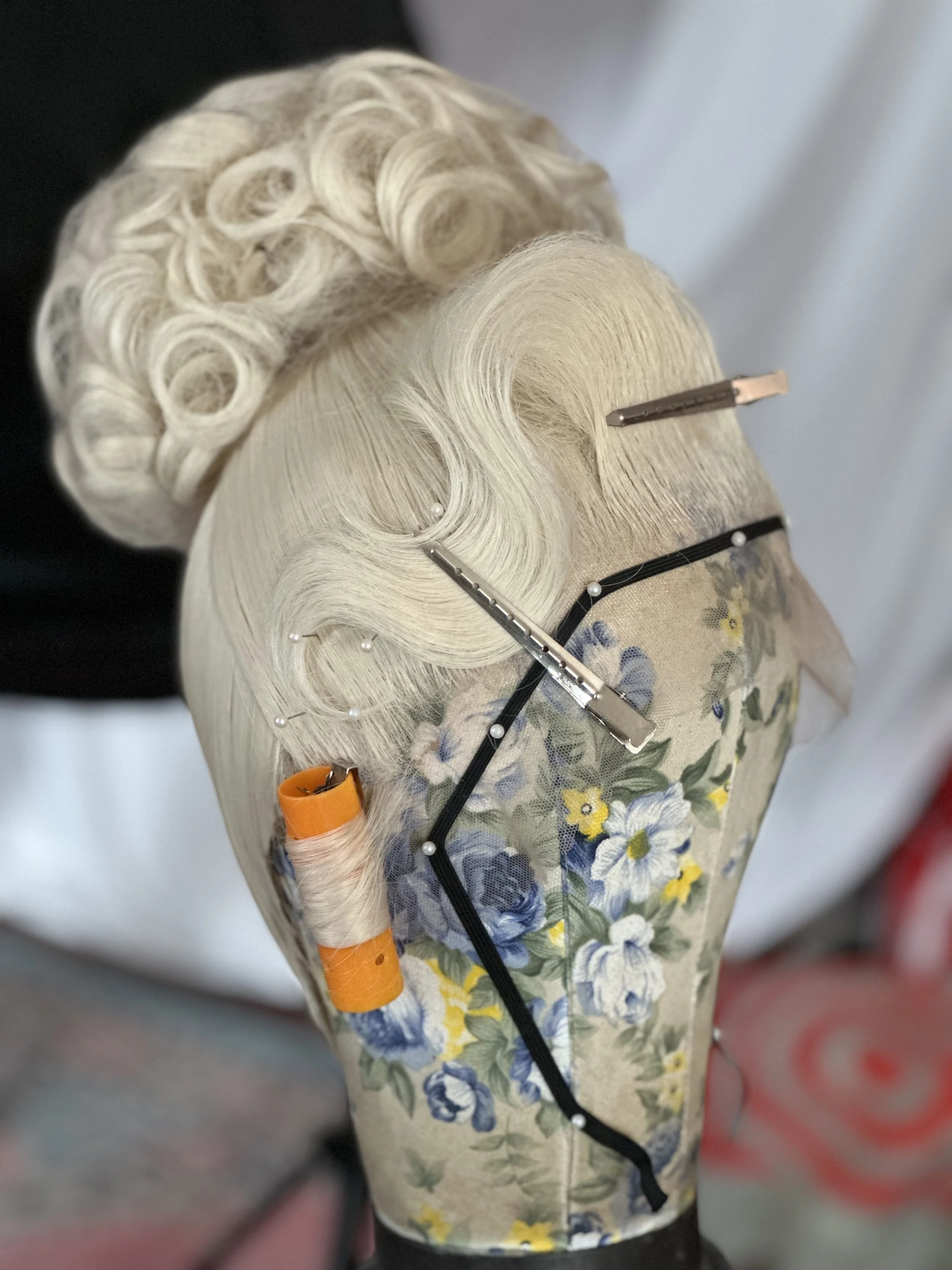 Close-up of a mannequin head with synthetic hair styled in vintage curls, fastened with hairpins, wearing a floral blouse with yellow, blue, and white flowers, and an orange hair roller visible at the bottom.