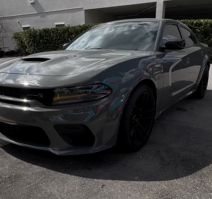 A gray Dodge Charger parked in a lot, with a building and bushes in the background.