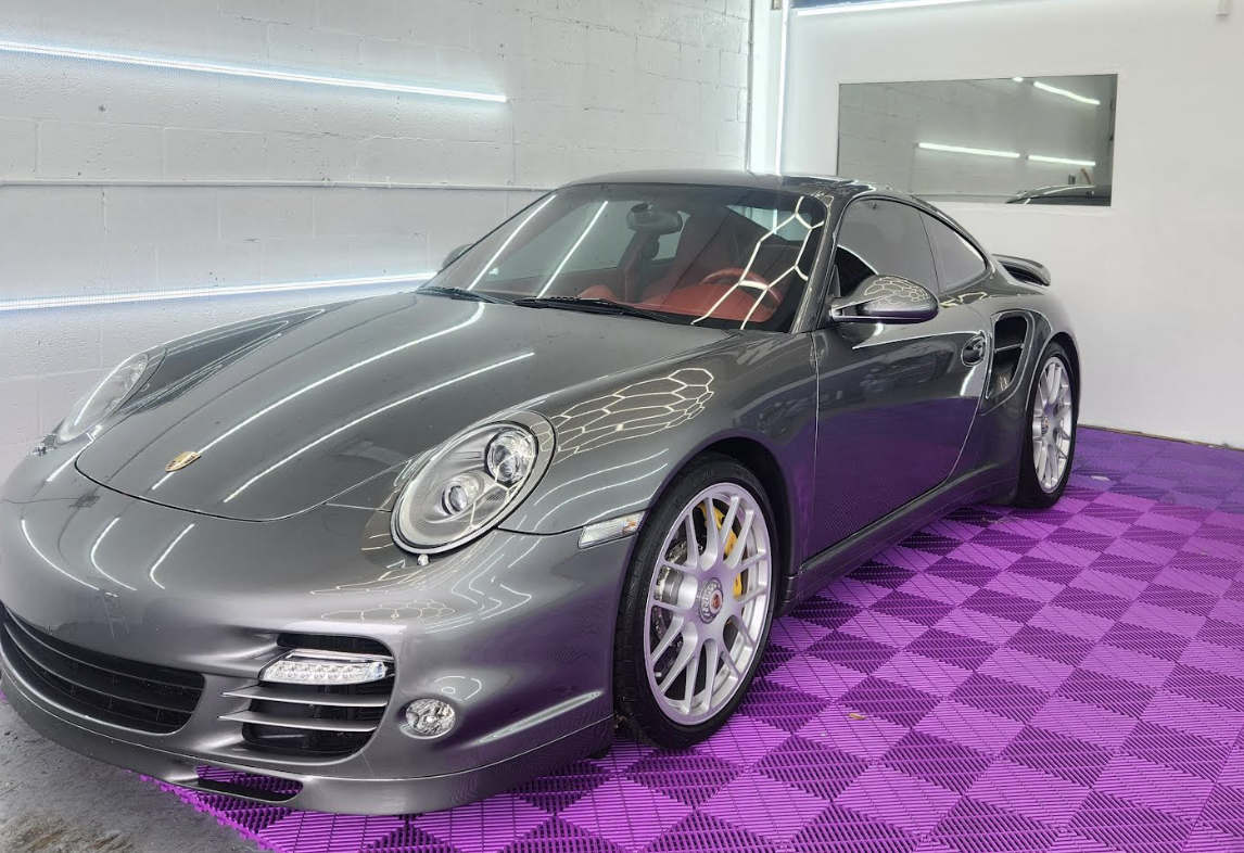 A silver Porsche sports car inside a showroom with purple checkered flooring and white walls, reflecting ceiling lights.