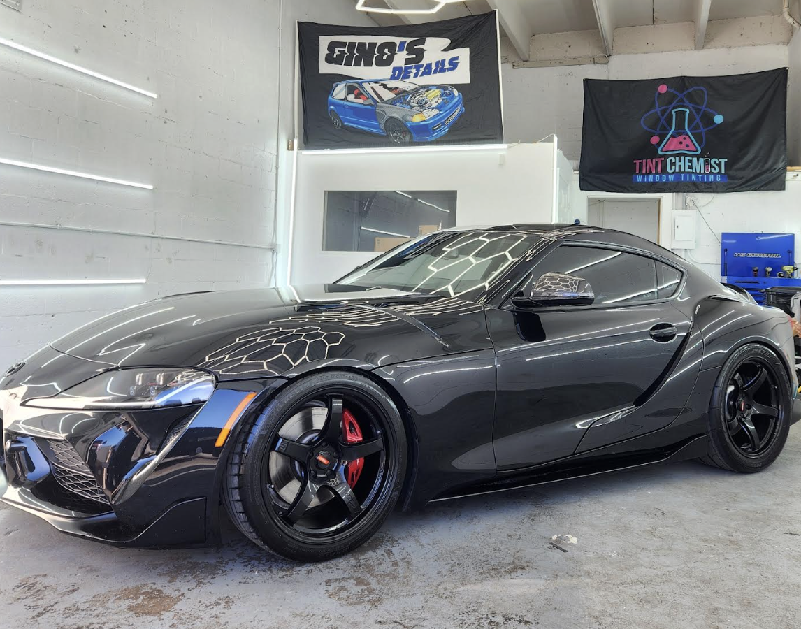A black sports car, likely a Toyota Supra, parked inside a detailing shop with banners on the wall reading 'S-ONE DETAILS' and 'TINT CHEMIST WINDOW TINTING.' The car has black wheels and red brake calipers.