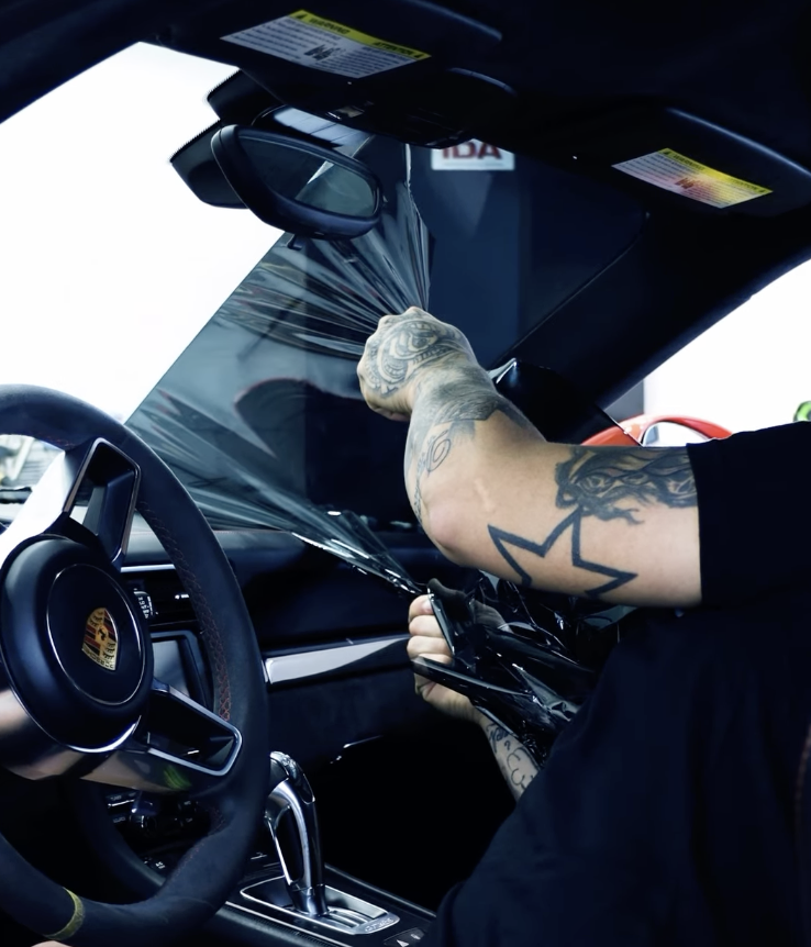 Person applying a tinted window film to the inside of a car windshield from the driver's side, with a Porsche steering wheel visible.