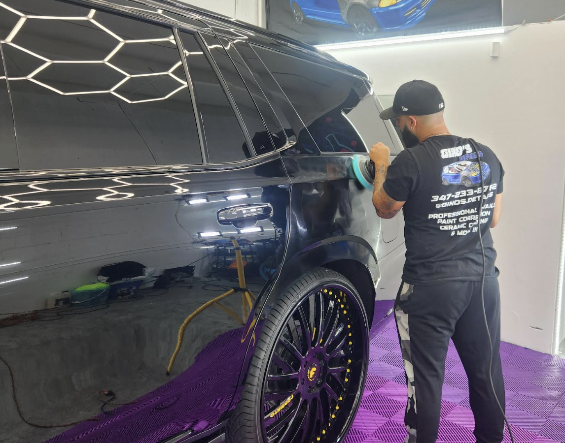 A man polishing the side of a black SUV inside a garage. The SUV has large black wheels with purple accents. There is a poster or picture of a blue car on the wall in the background.