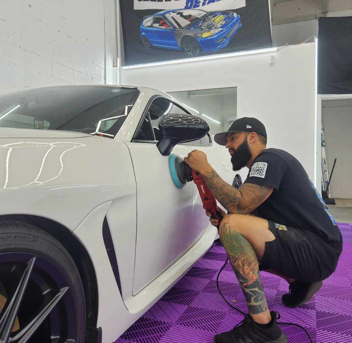 A man with tattoos, wearing a black cap and black t-shirt, is polishing a white sports car with a power buffer in a garage. There is a poster of a blue racing car on the wall above.