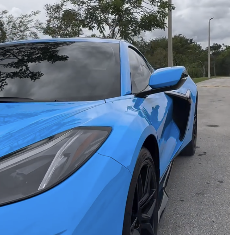 A close-up of a blue sports car parked on a street, showing the front and side, with the clouded sky and some trees in the background.