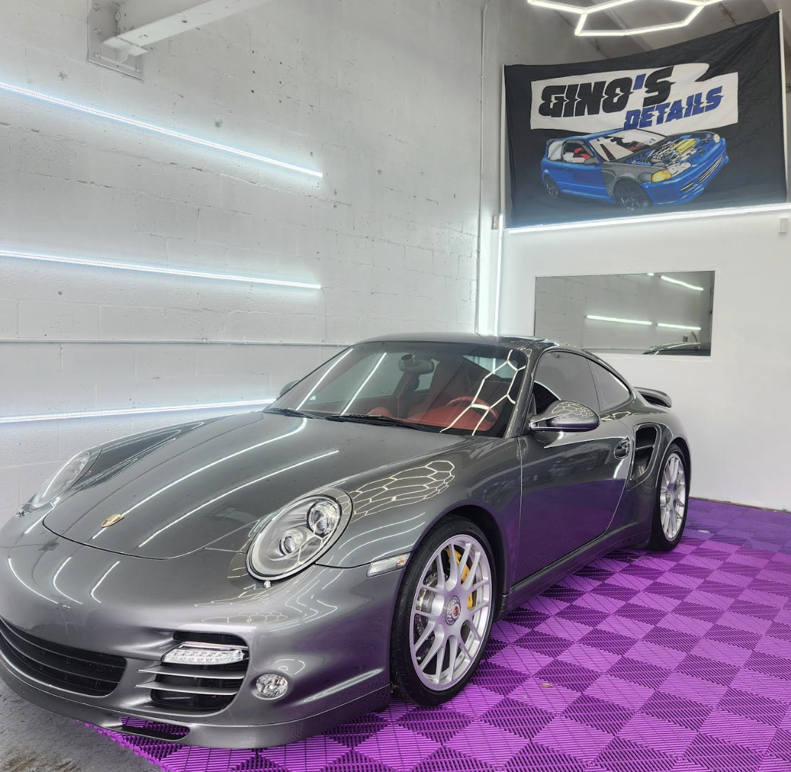 A gray Porsche sports car parked inside a showroom with modern white walls and neon lighting. There is a black and blue banner with a car illustration and the text 'S & B R's Details' hanging on the wall. The floor features purple checkered tiles.
