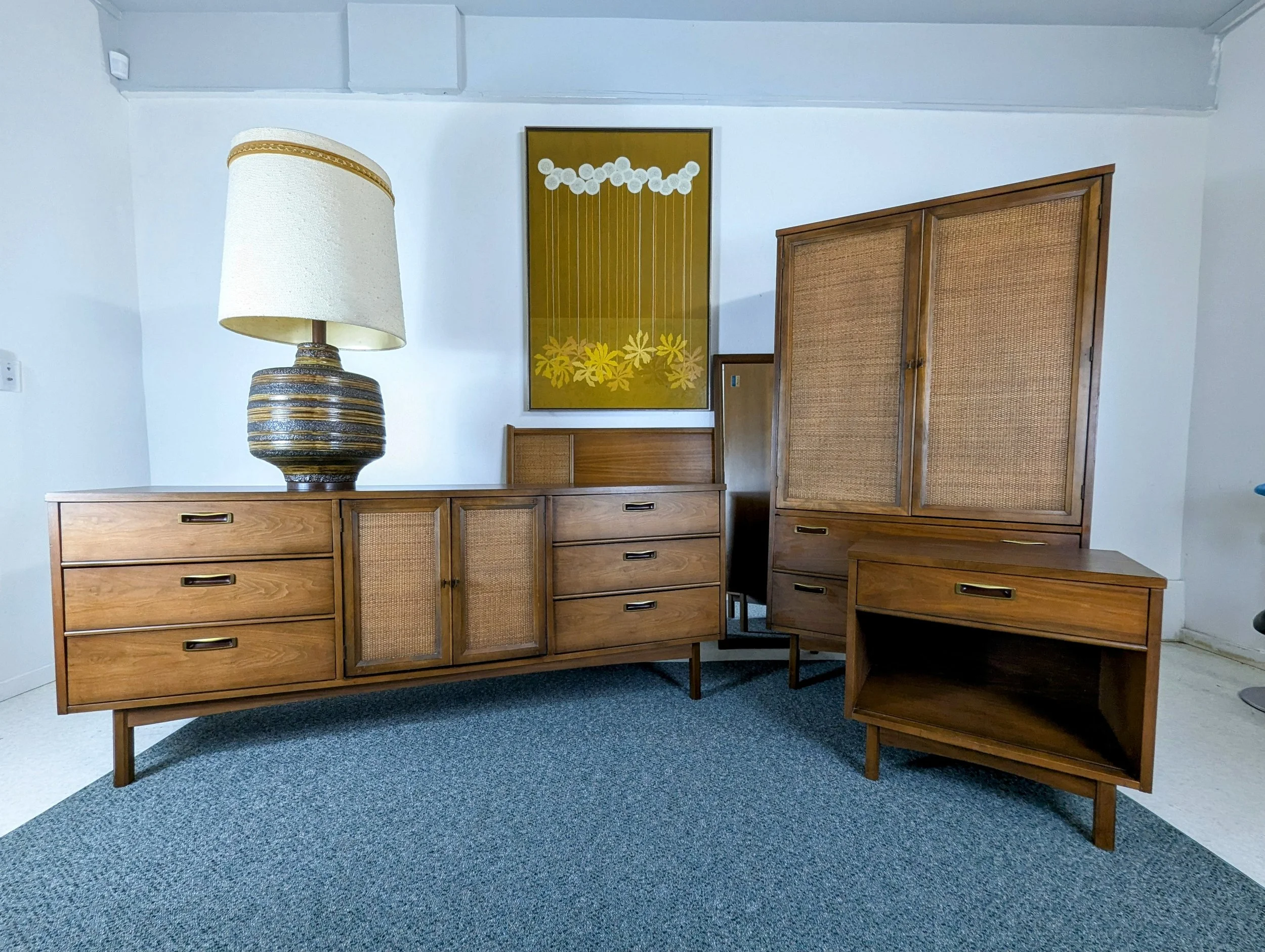 Mid-century modern wooden furniture set with a large table lamp, a framed floral artwork, several cabinets with rattan panel doors, and a small open shelf, all arranged on a blue carpet in a room with white walls.
