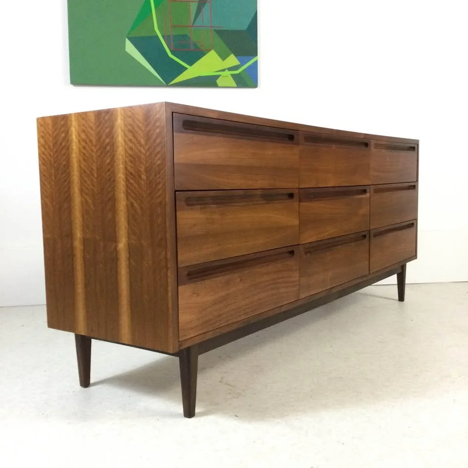 A mid-century modern wooden sideboard with six drawers, standing on four tapered legs, set against a plain wall with a colorful abstract painting above it.