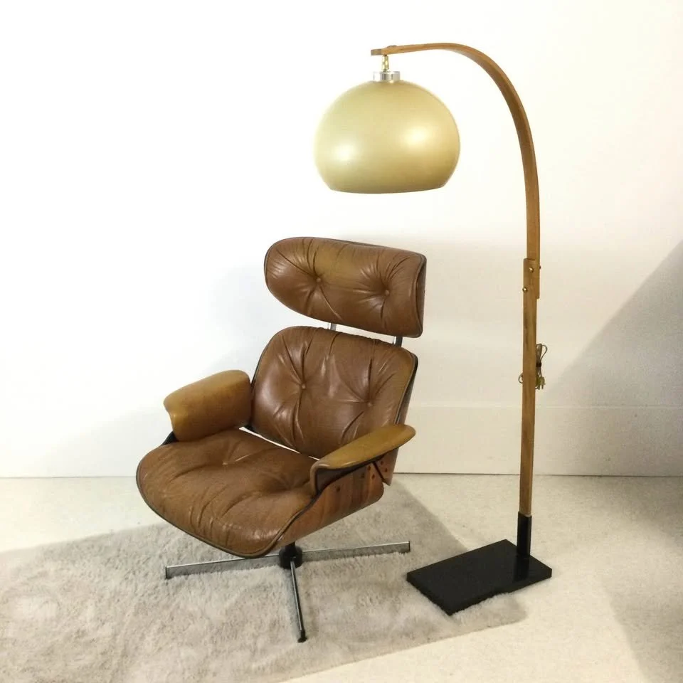 A vintage brown leather lounge chair with a matching headrest and an attached slide armrest, positioned next to a tall wooden floor lamp with a large, cream-colored, rounded lampshade, set against a plain white wall and light-colored carpet.