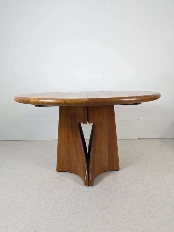 A round wooden table with a sculptural base, placed against a plain white wall.
