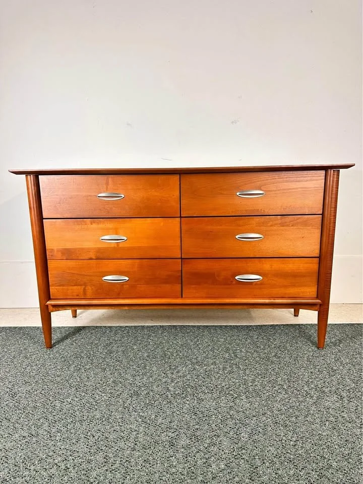 A mid-century modern wooden dresser with six drawers and metal handles, standing against a white wall on a carpeted floor.