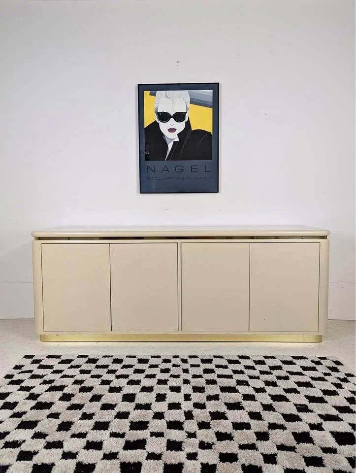 A framed stylish poster of a woman with blonde hair, sunglasses, and dark lipstick hangs on a plain white wall. Below the poster is a beige sideboard, and in front is a black and white patterned rug.
