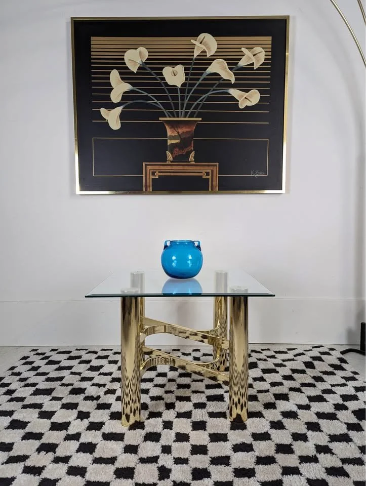 Decorative interior scene featuring a glass-top table with gold-colored legs, a blue vase in the center, and two small white candles. A framed art piece of a vase with stylized flowers hangs on the wall above, and there is a patterned black and white