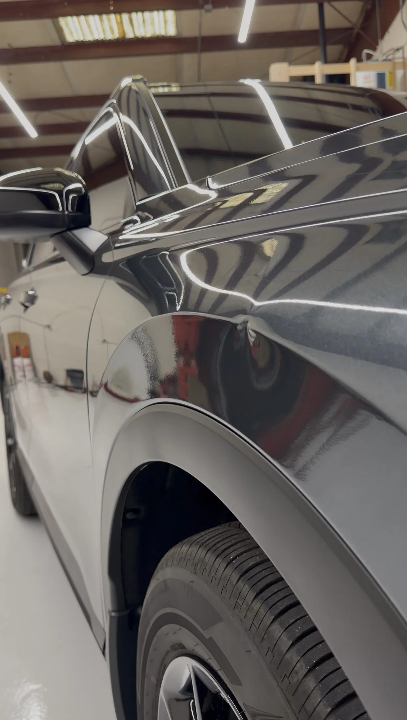 A shiny black and silver car parked inside a garage, with reflections of the garage ceiling and fluorescent lights on its surface, showing part of the front and side of the vehicle.
