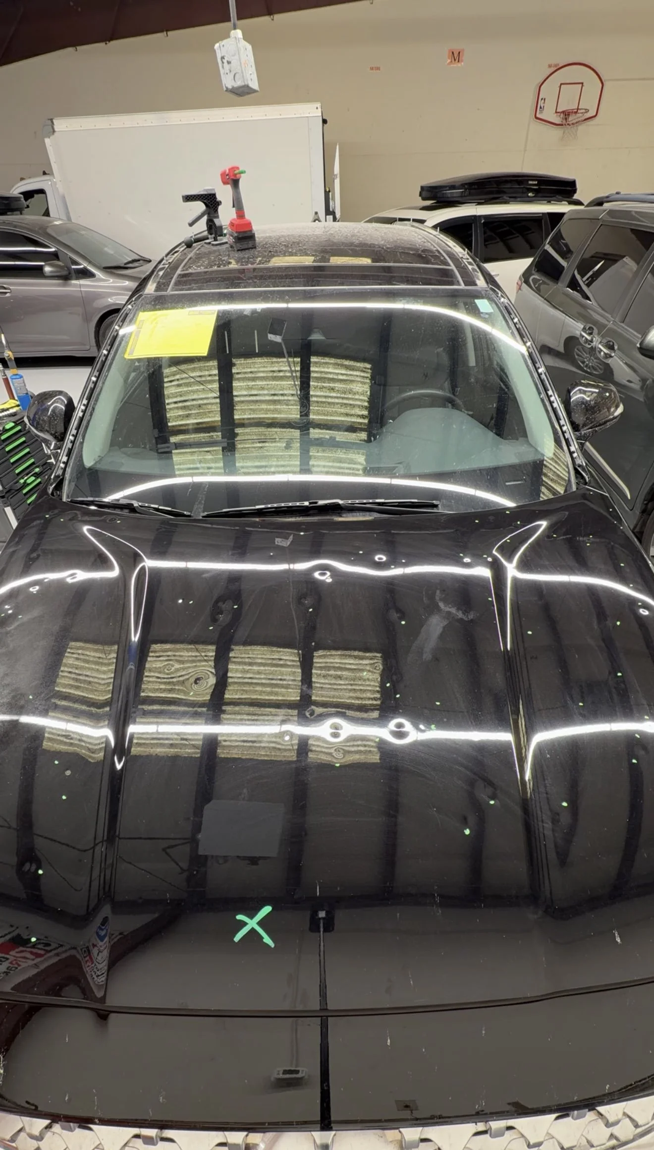 A black SUV inside a garage or workshop, with a yellow paper or sign visible on the windshield, and hail damage on the hood.