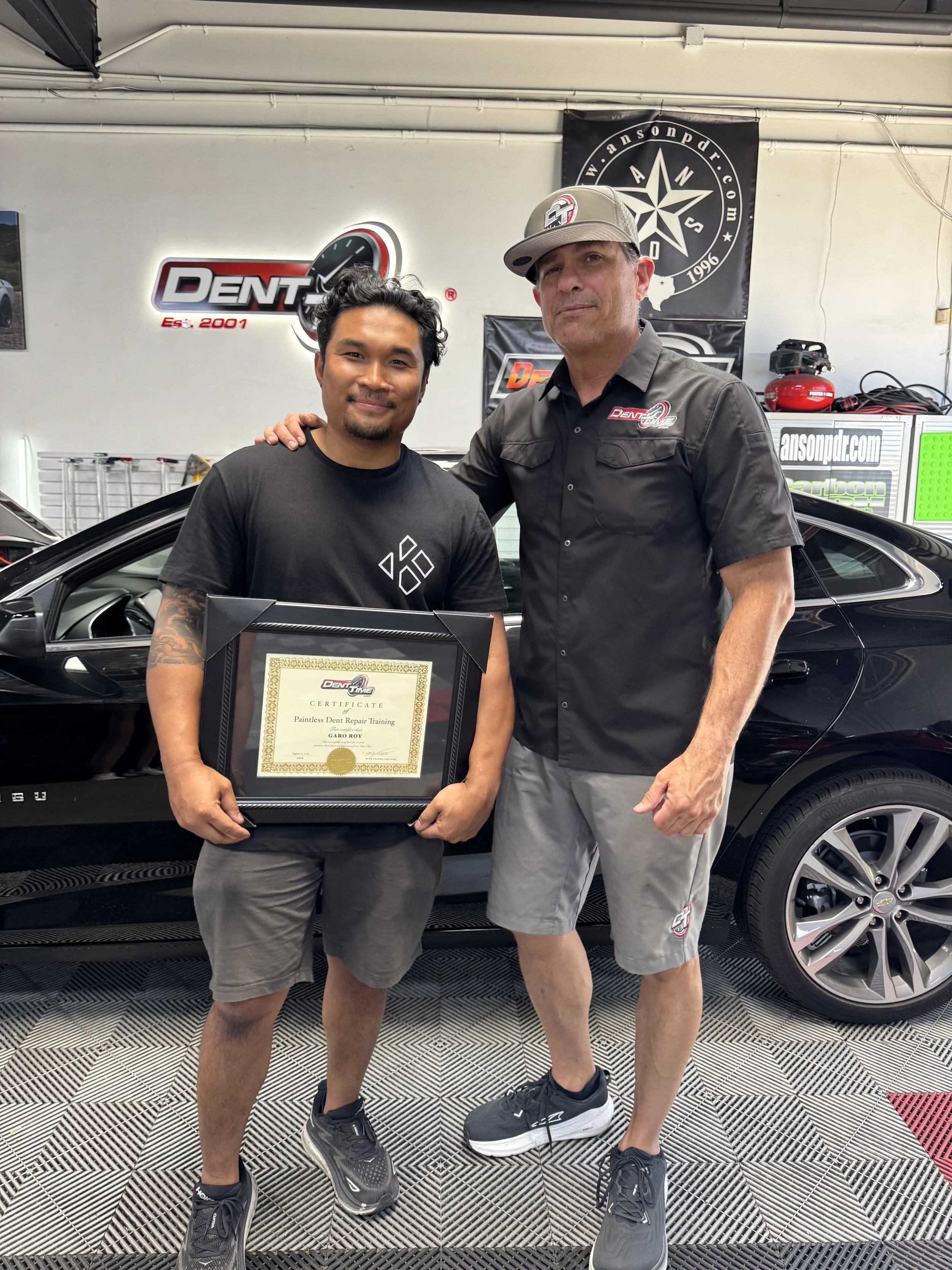 Two men standing in a garage, one holding a certificate, with a black sports car behind them. The man on the left is wearing a black t-shirt and gray shorts, while the man on the right is wearing a black short-sleeve shirt, gray shorts, and a gray baseball cap. There are automotive banners and equipment visible in the background.