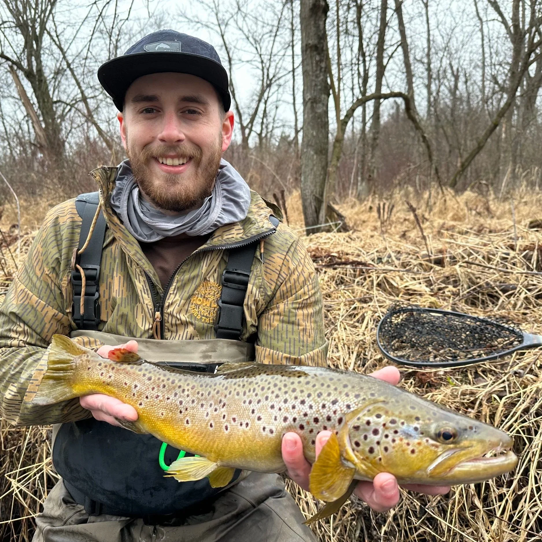 A smiling man in outdoor gear holding a large brown trout with spots, standing in a grassy, wooded area.