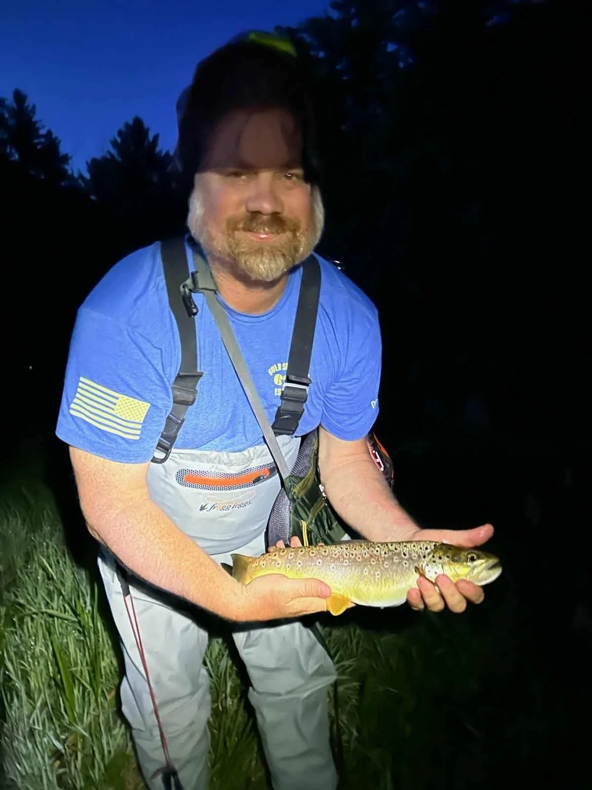 A man with a beard, wearing a blue t-shirt and fishing gear, holding a brown and yellow spotted fish at dusk outdoors.