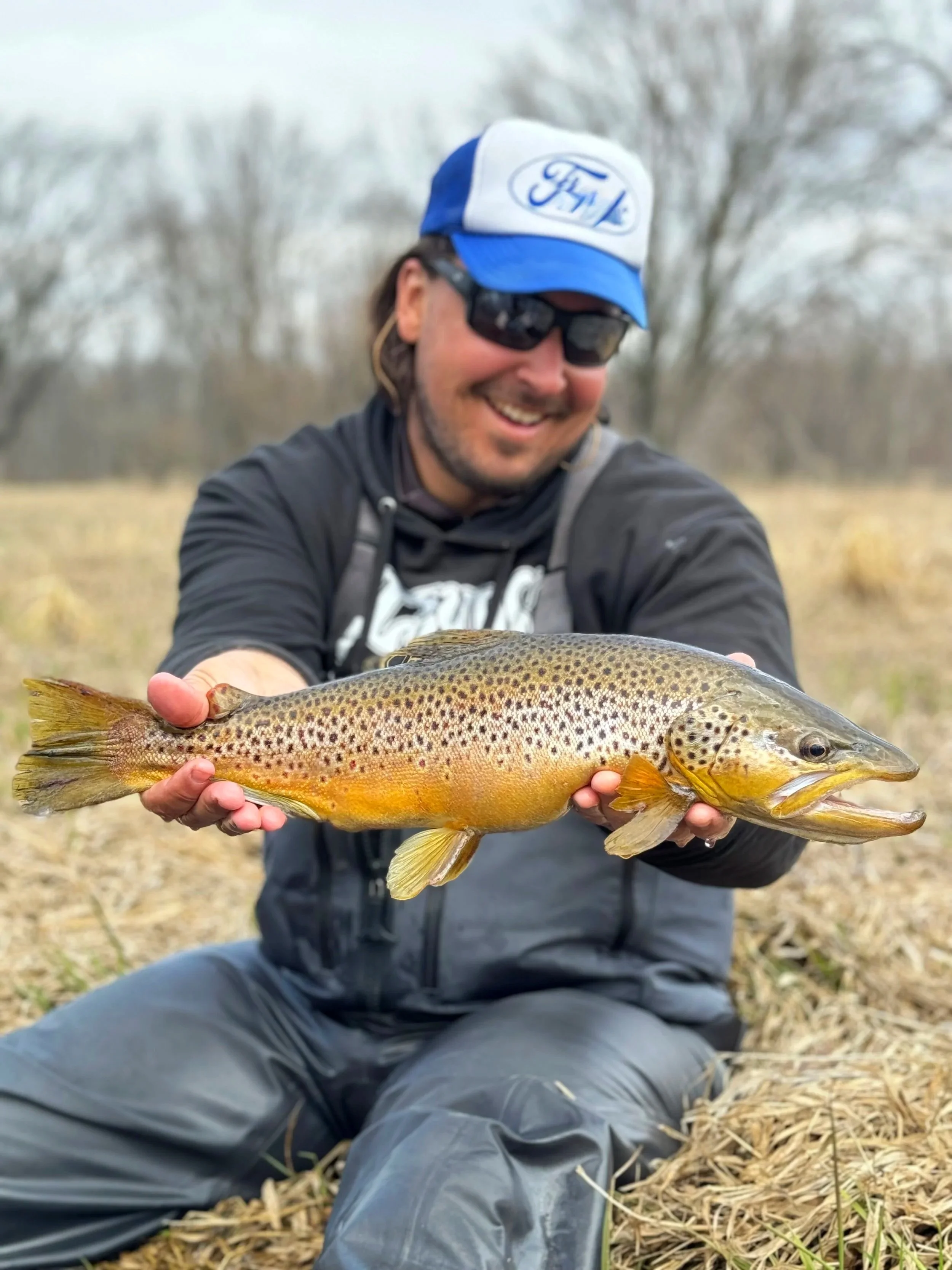 A smiling man wearing sunglasses and a blue and white Ford hat, sitting outdoors on dry grass with trees in the background, holding a large brown trout fish with distinctive spots and coloration.