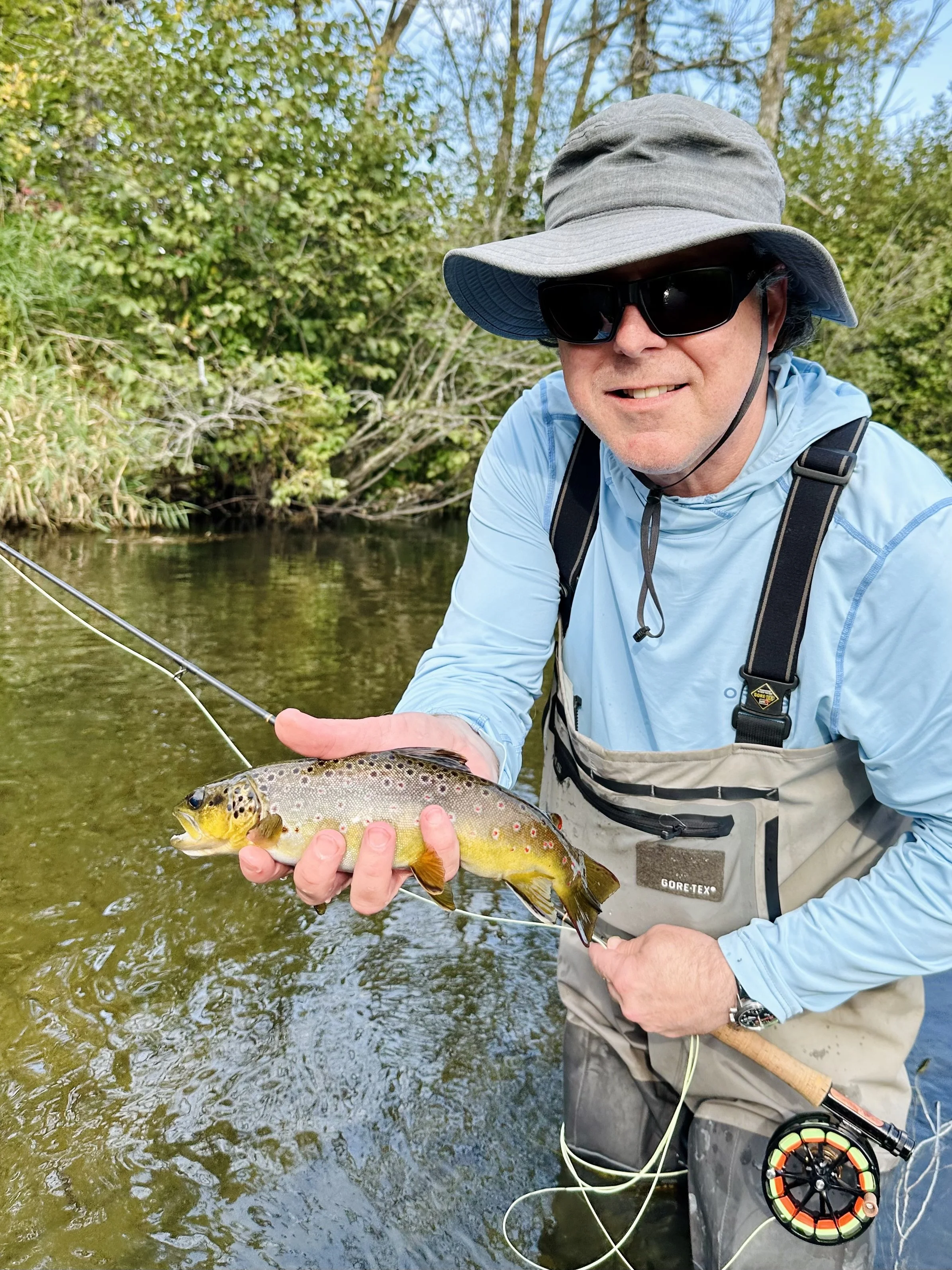 Man wearing sunglasses, a large hat, and fishing gear, holding a caught rainbow trout over a river with green foliage in the background.