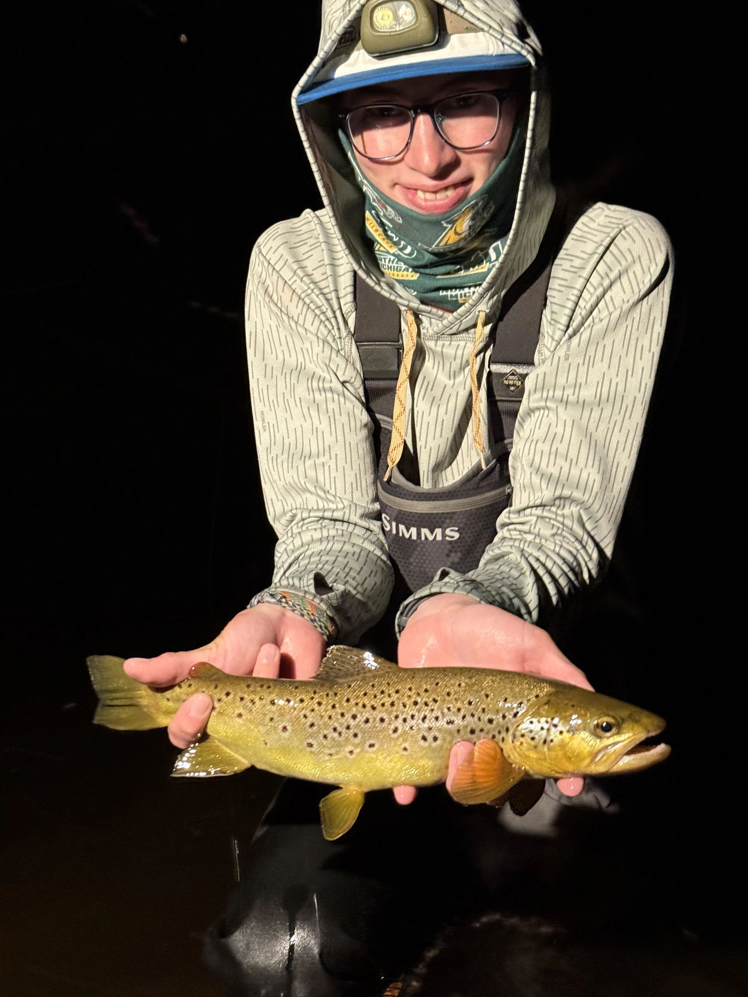 Person wearing fishing gear holding a brown trout fish at night.