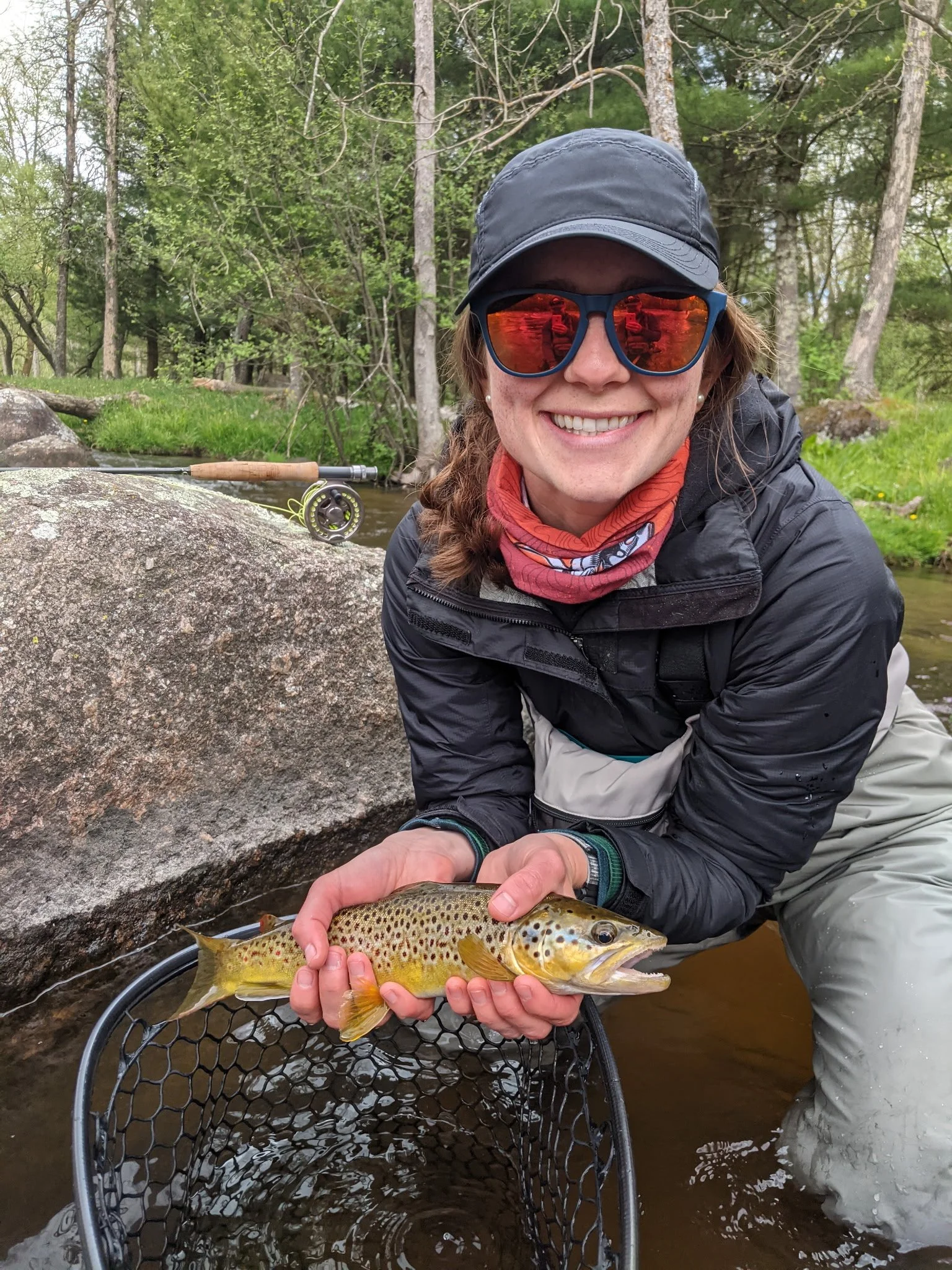 A woman wearing sunglasses, a black cap, and outdoor clothing is kneeling by a stream, smiling and holding a caught trout over a fishing net.