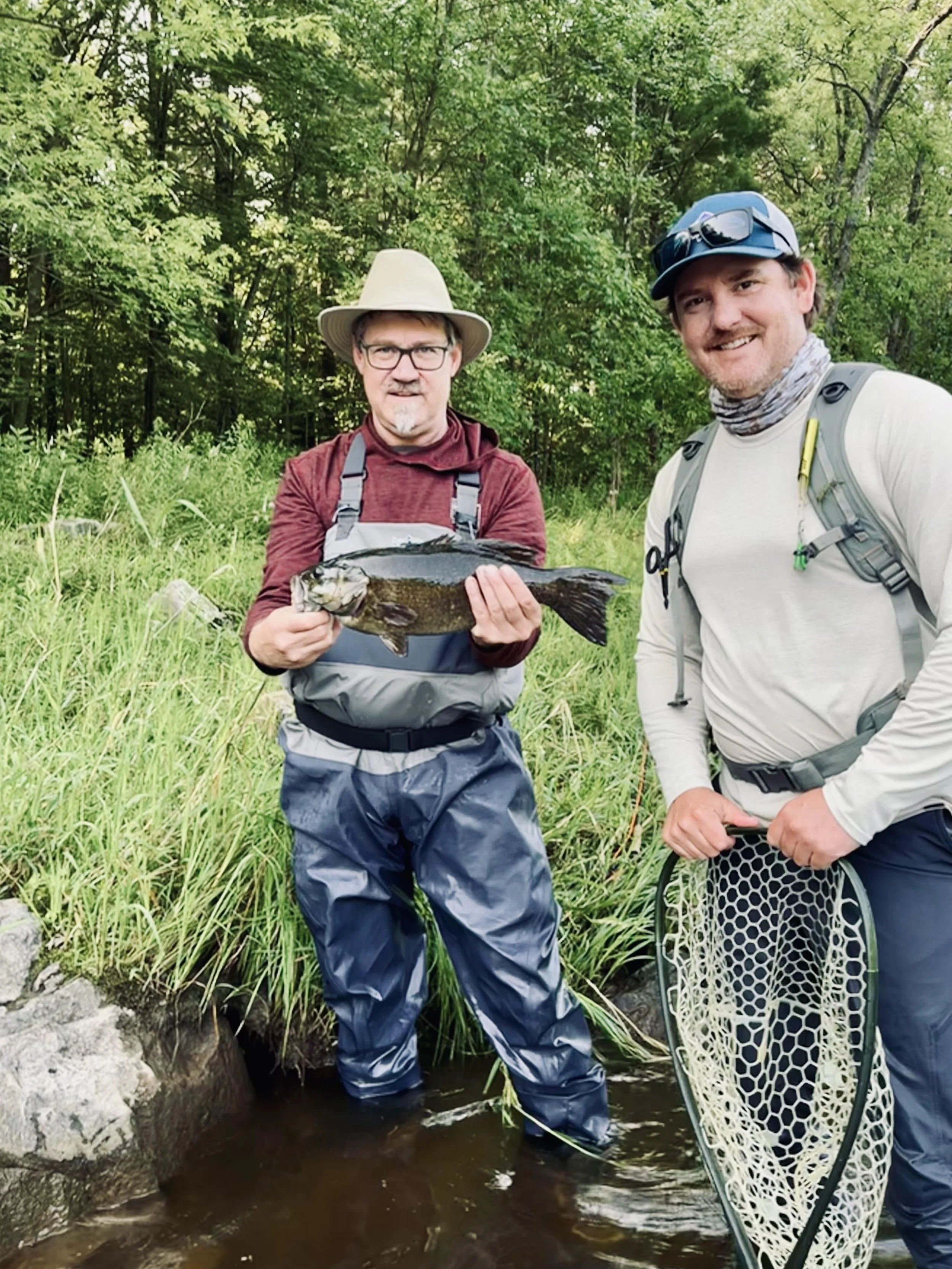 Two men outdoors, one holding a fish, beside a stream, with trees and greenery in the background.