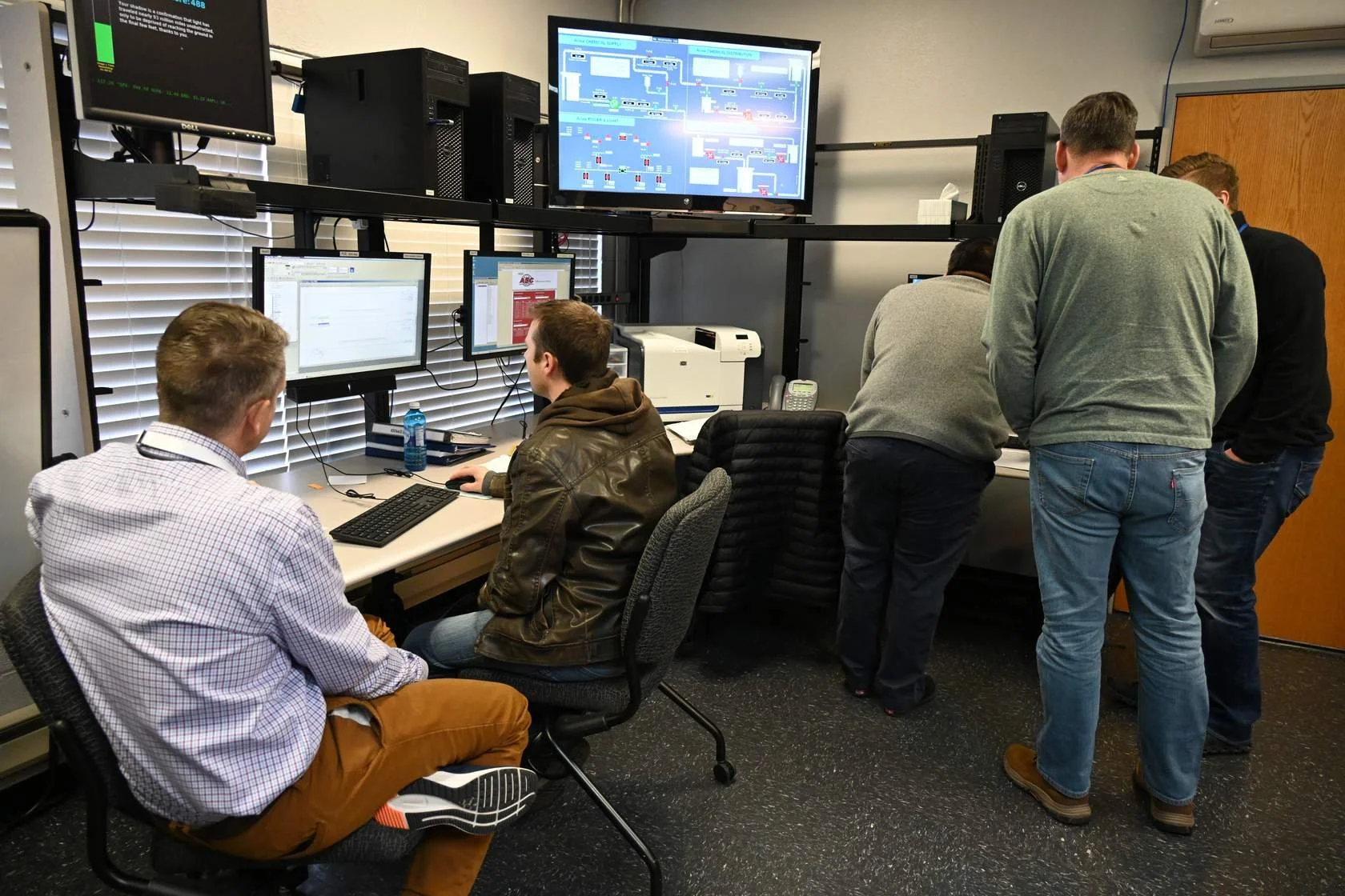 Group of five people working in a control room with computer monitors and a large screen displaying a flowchart or system diagram.