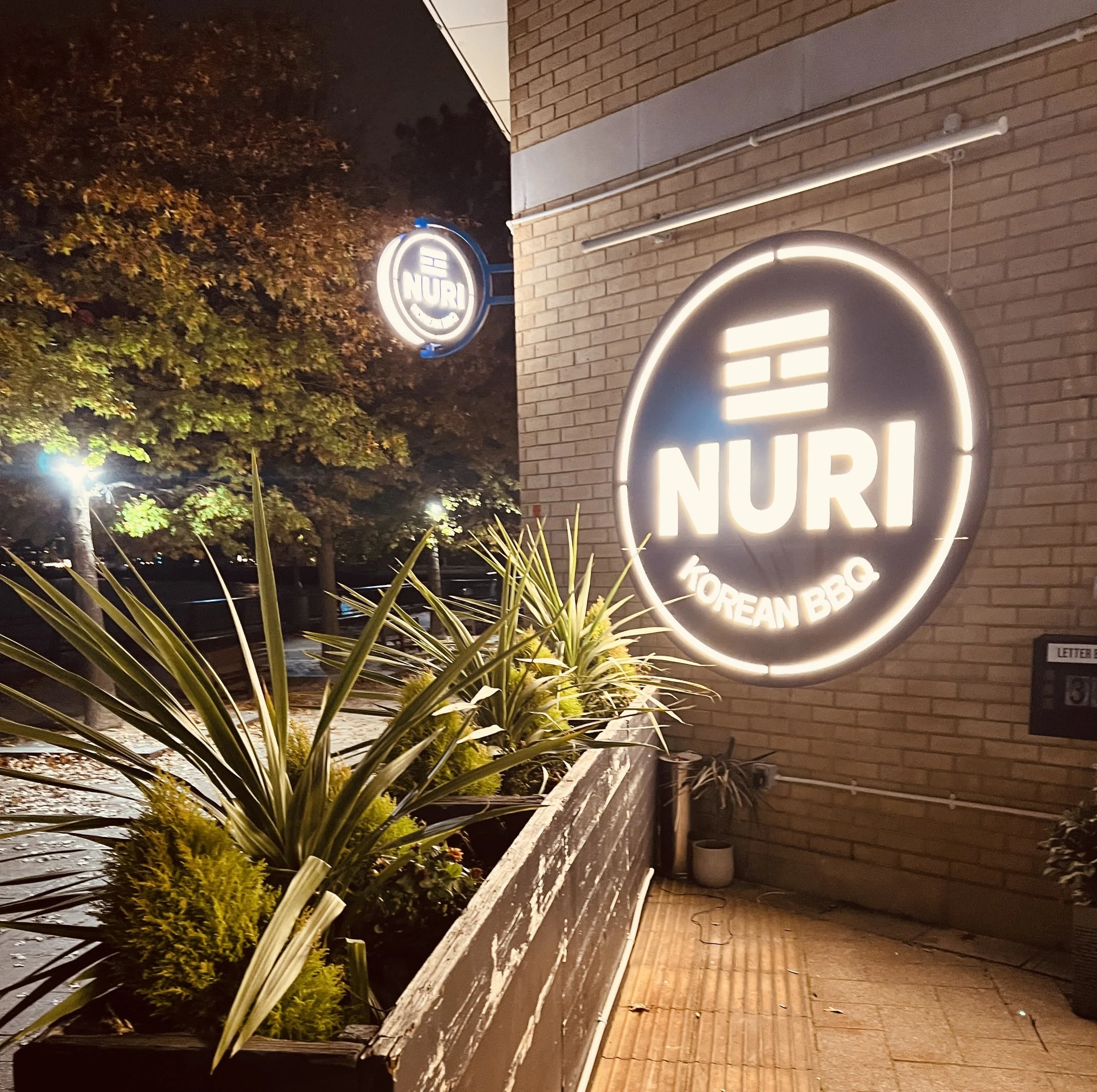 A brightly lit circular sign outside a restaurant reads 'NURI KOREAN BBQ'. The sign is mounted on a brick wall, with a planter box containing various green plants and a small potted plant underneath. There are trees and streetlights in the background