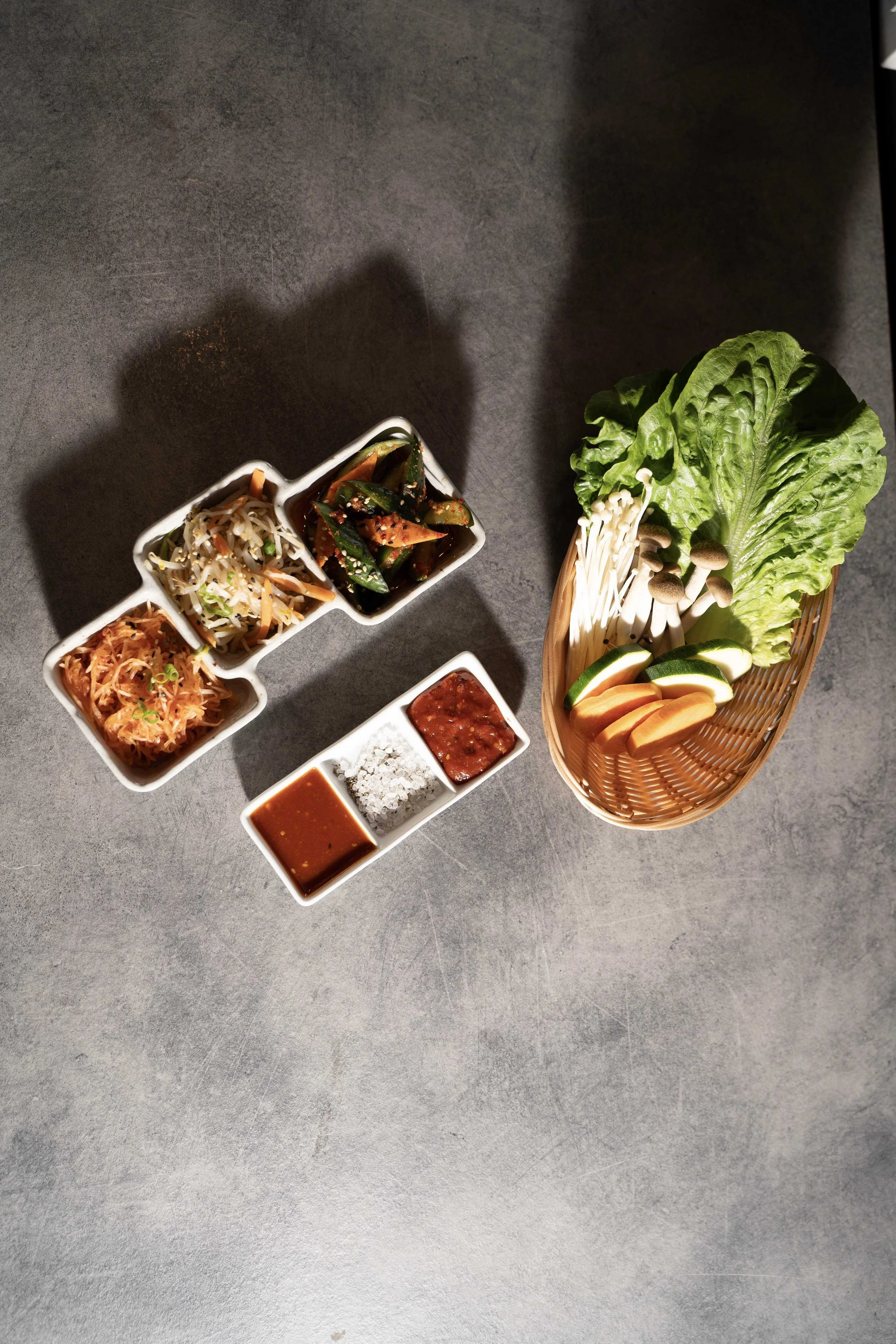 Assorted Korean banchan dishes with fresh lettuce and vegetables on a gray surface.