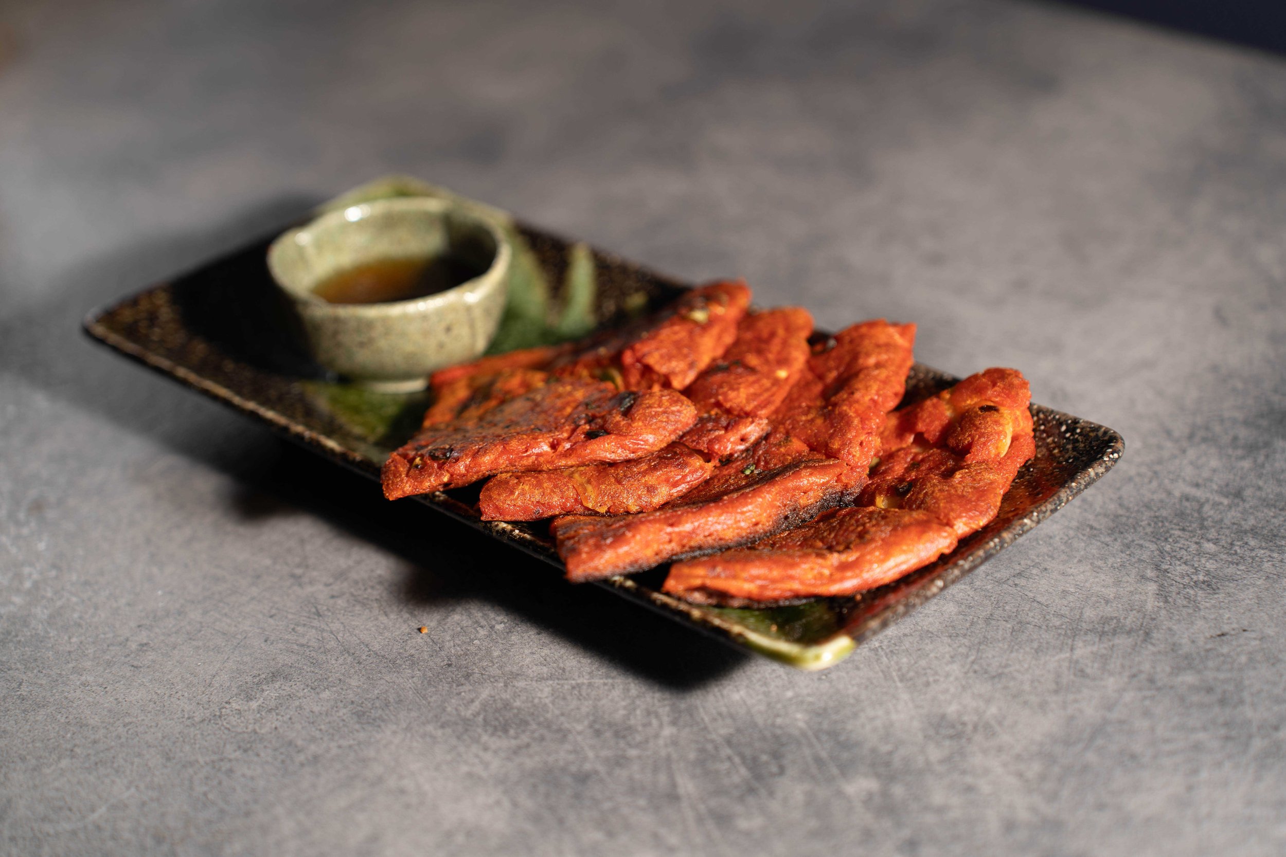 A rectangular black plate with slices of spicy, grilled meat garnished with sauce in a small bowl on the side, set on a gray surface.