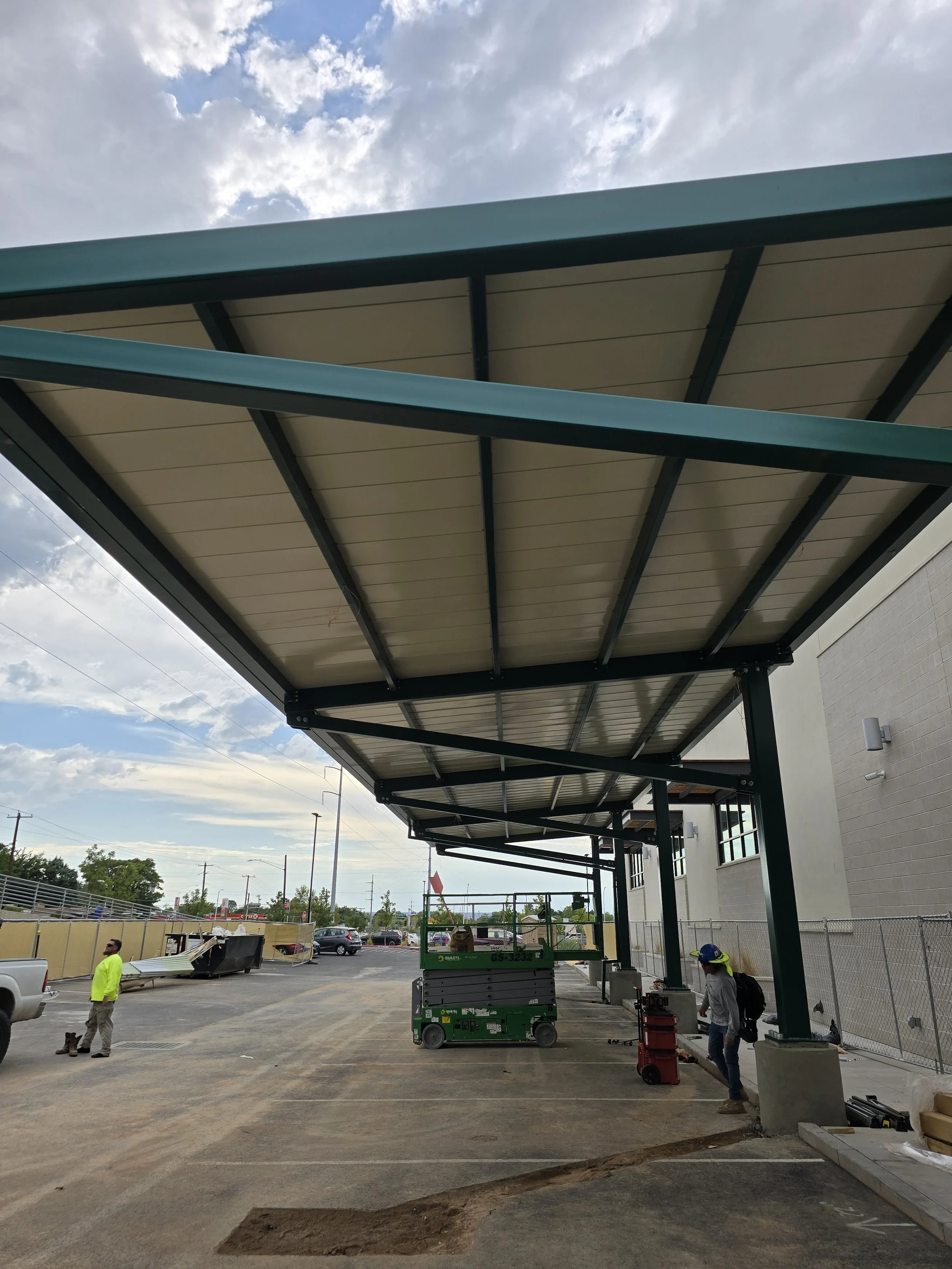 Construction workers installing a large green metal awning on a building in a parking lot with vehicles, a fence, and utility poles visible in the background.