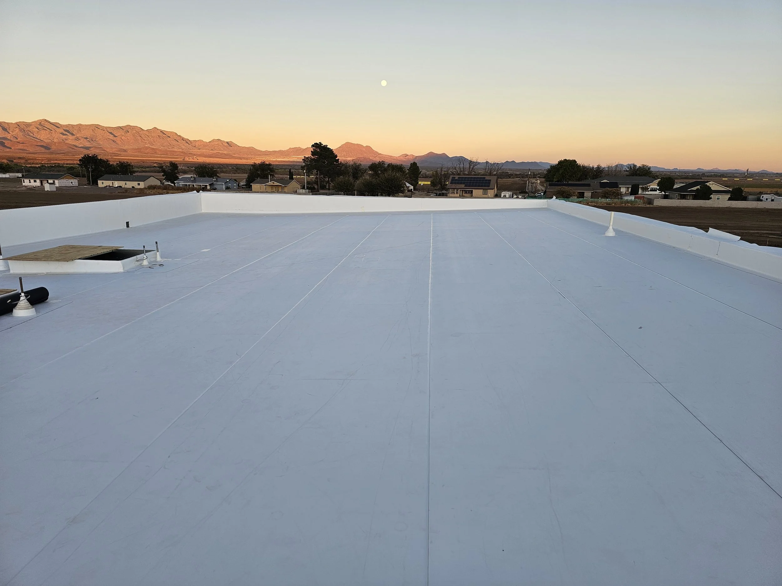 View of a flat rooftop covered with white reflective material, with a small wooden platform and ventilation pipes. In the background, there are suburban houses, trees, mountains, and a clear sky with a visible moon.