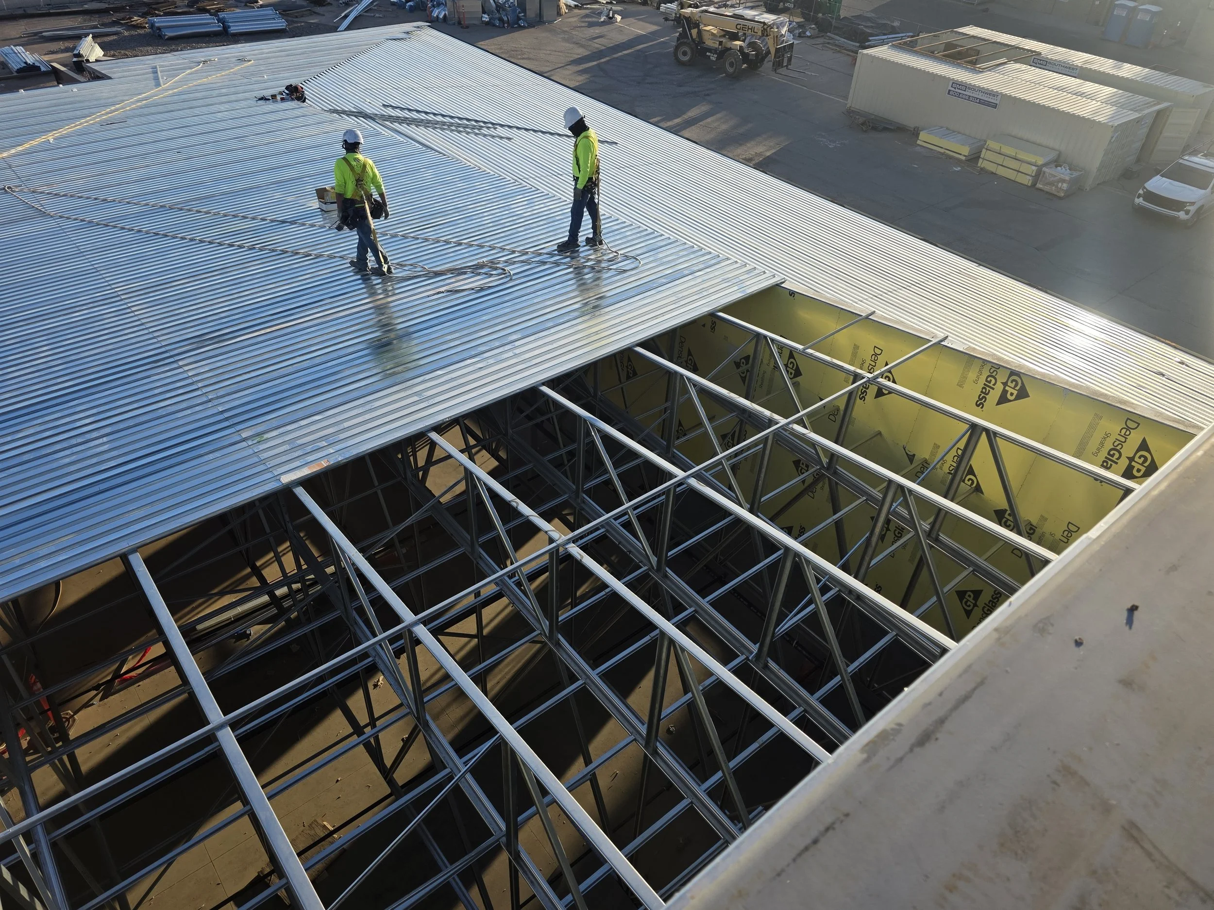 Two construction workers in yellow safety vests and white helmets installing a corrugated metal roof on a building, with an unfinished metal framework visible underneath.
