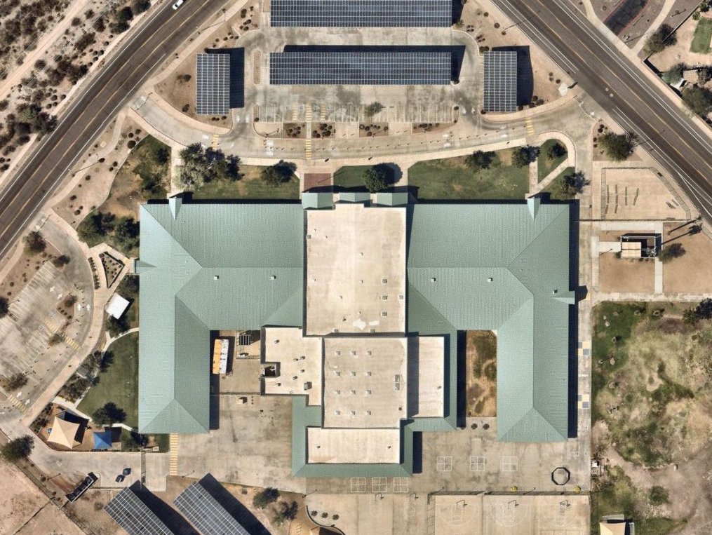 An aerial view of a large school building with a green roof, surrounded by playgrounds, parking lots, and solar panels, with highways nearby.