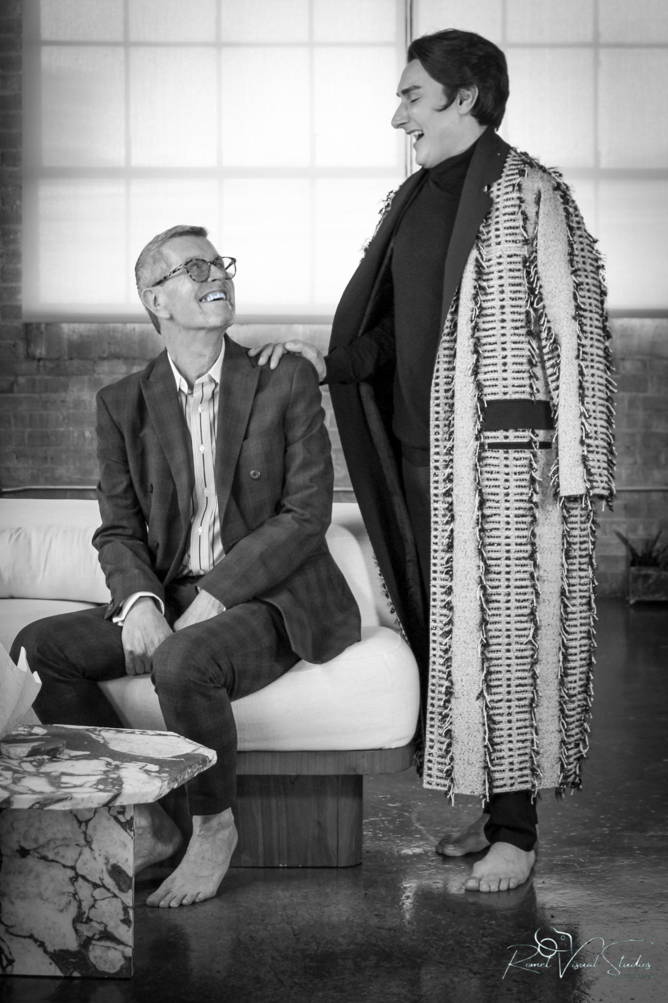 Two men having a conversation indoors, with one sitting on a sofa and the other standing next to him, both smiling and appearing happy.