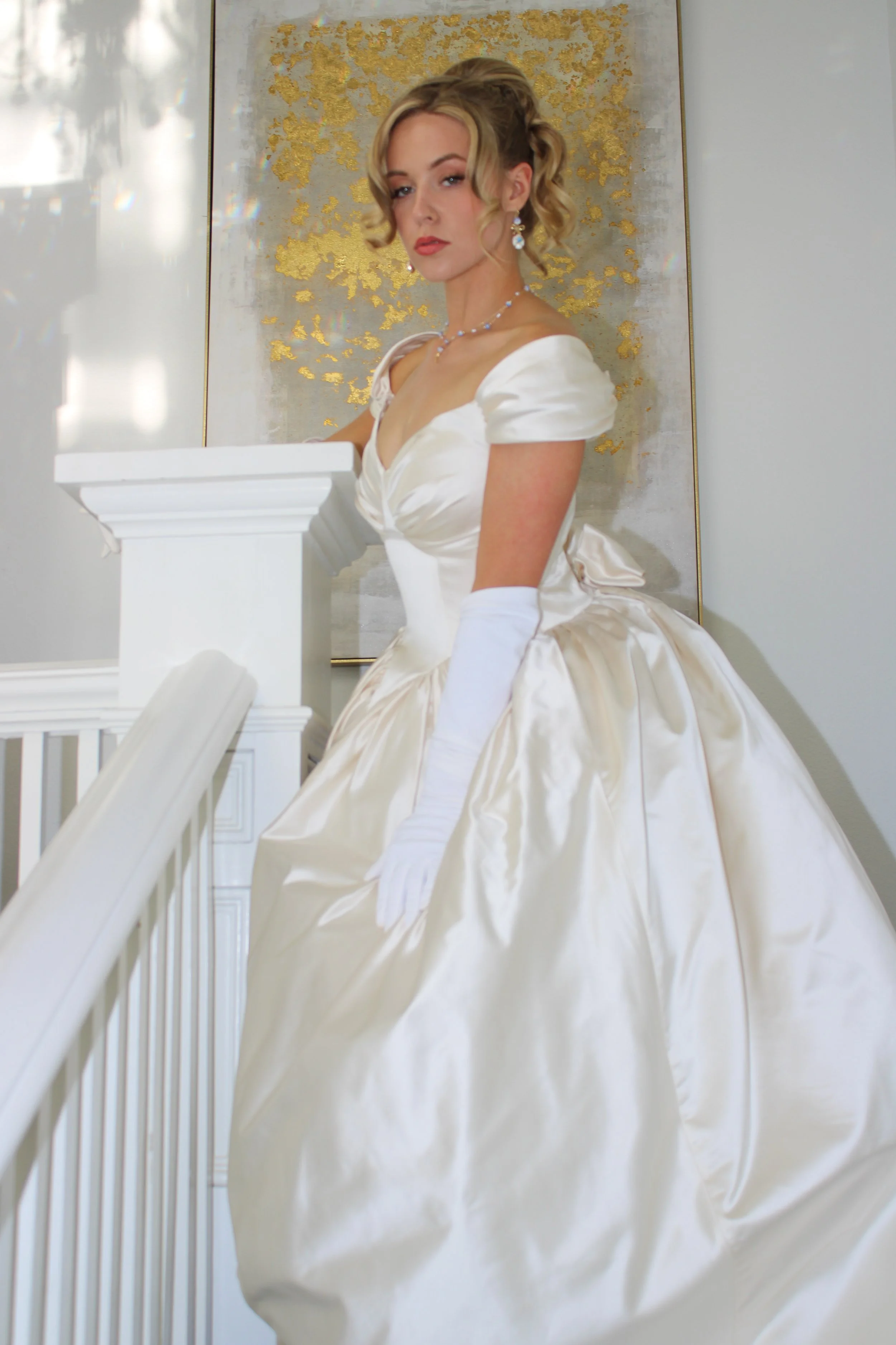 A woman in a vintage white wedding gown with gloves, posing on a staircase indoors.