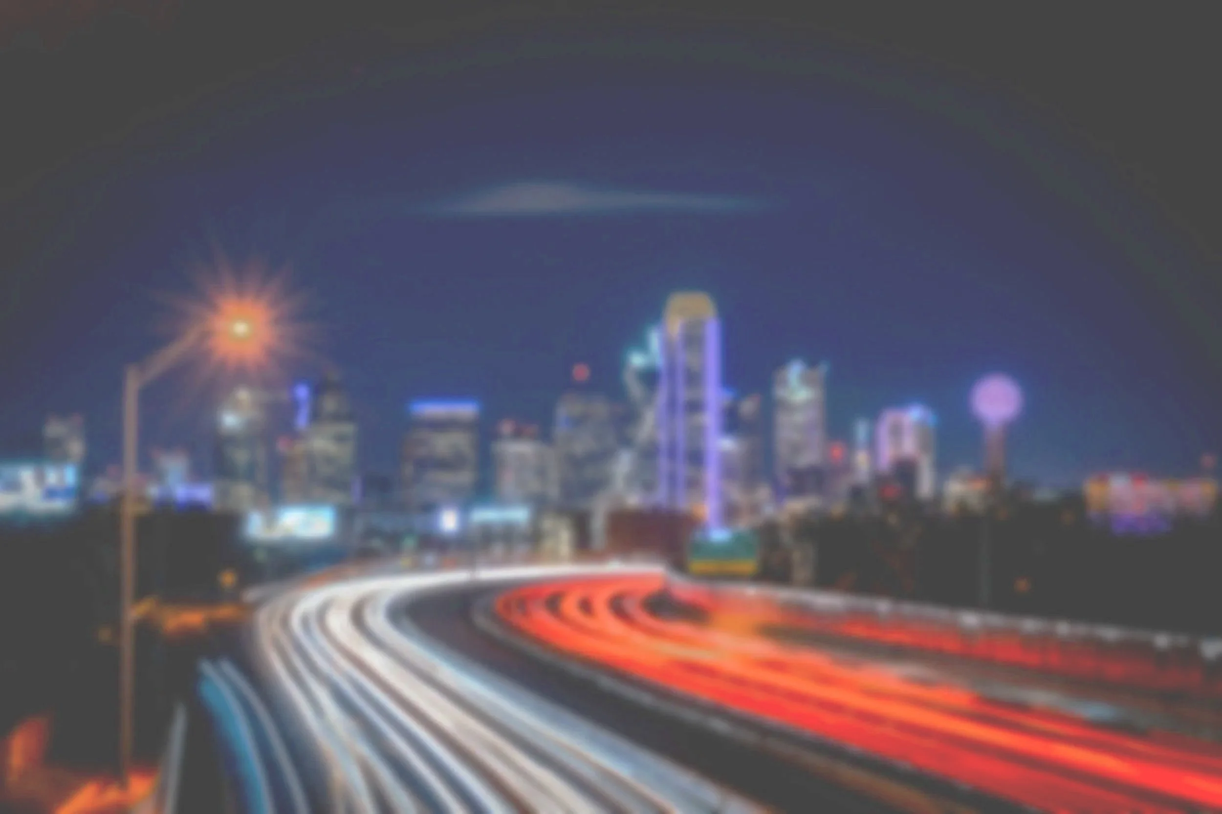 Blurred night cityscape with illuminated skyscrapers and streaks of red and white lights from moving vehicles on a highway.