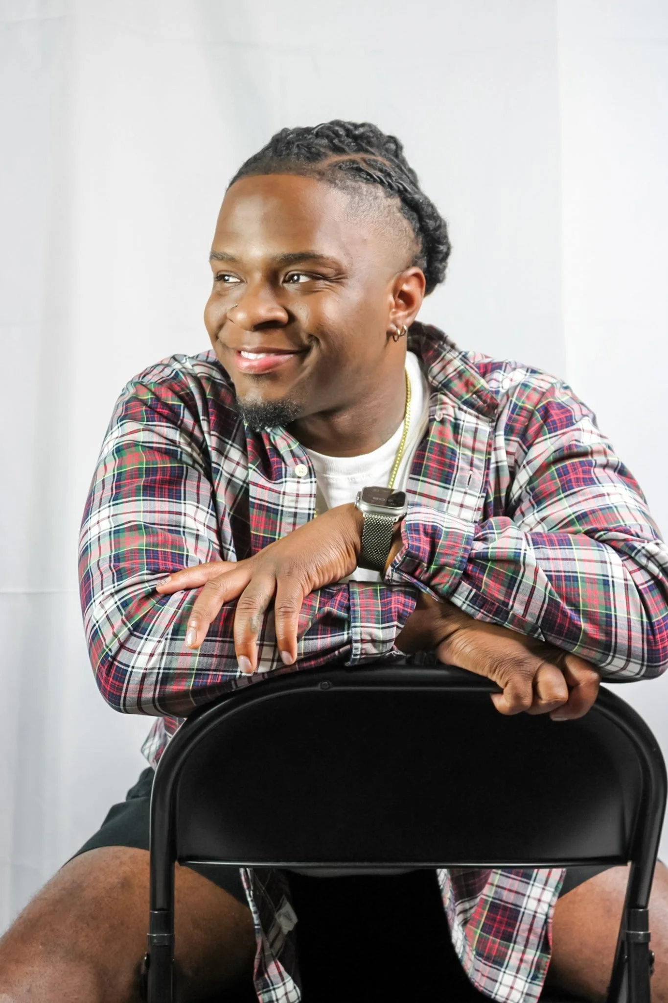 A young man smiling brightly, sitting and leaning on a black chair, with his arms crossed over the top of the chair. He has short, braided hair, earrings, and is wearing a plaid shirt, a white t-shirt, a smartwatch, and a necklace. The background is plain white.