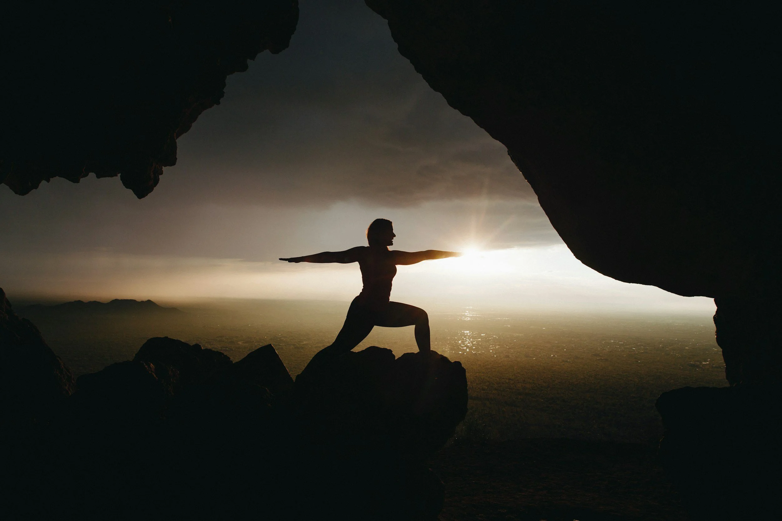 A person practicing yoga in a yoga pose, balancing with one knee on a rock, arms extended, between two large rock formations at sunset or sunrise with the ocean and mountains in the background.