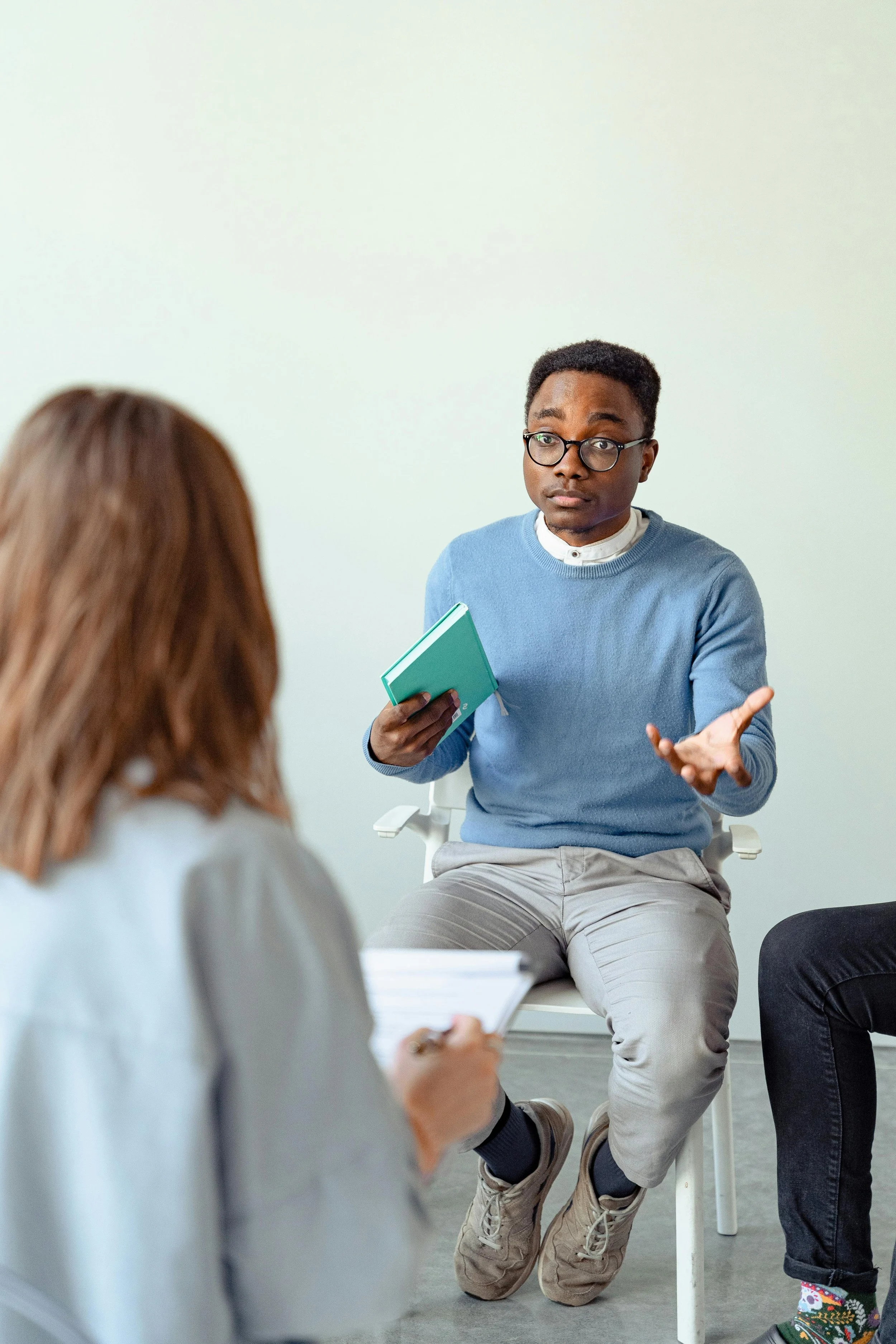 Man in glasses wearing a blue sweater holding a notebook talking to a woman with long red hair who is taking notes in a notebook, in a room with white wall.