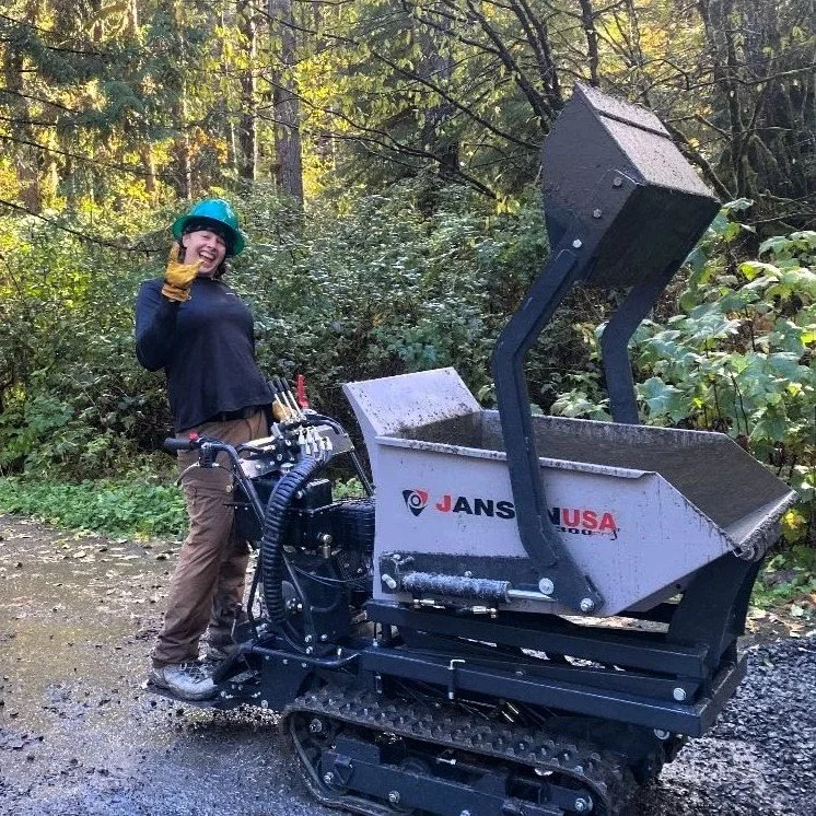 Young woman with hard hat driving track dumper, smiling outdoors with trees and greenery in the background.