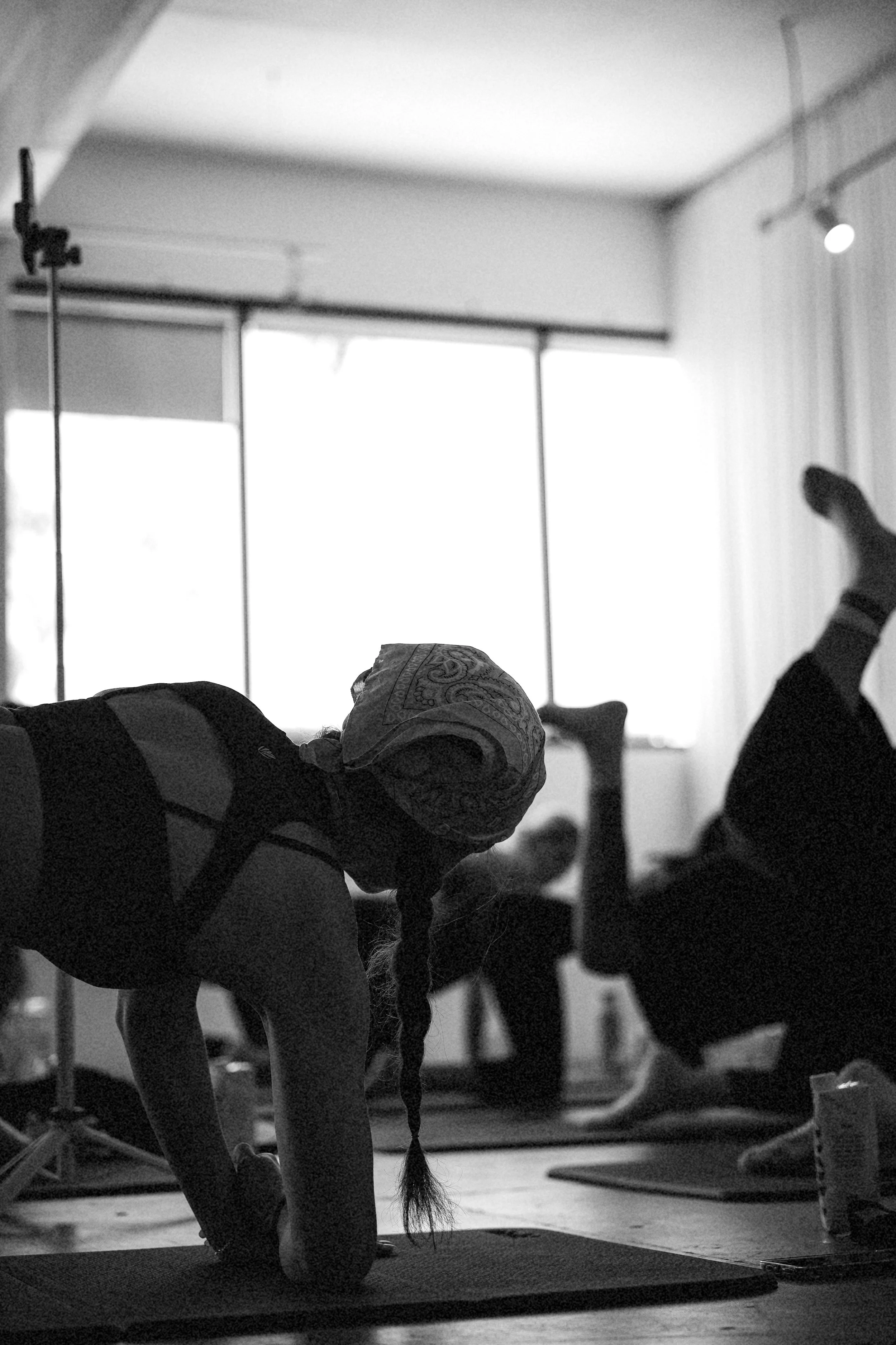 A person practicing yoga in a studio, in a forward bend position on a yoga mat, with other individuals and yoga props visible in the background.