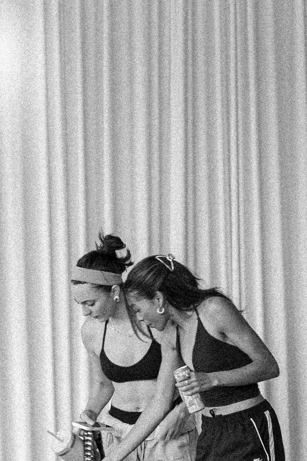 Two women dressed in black sports bras and pants, with one woman wearing a headband, are looking at something in their hands while standing close together in front of a curtain.