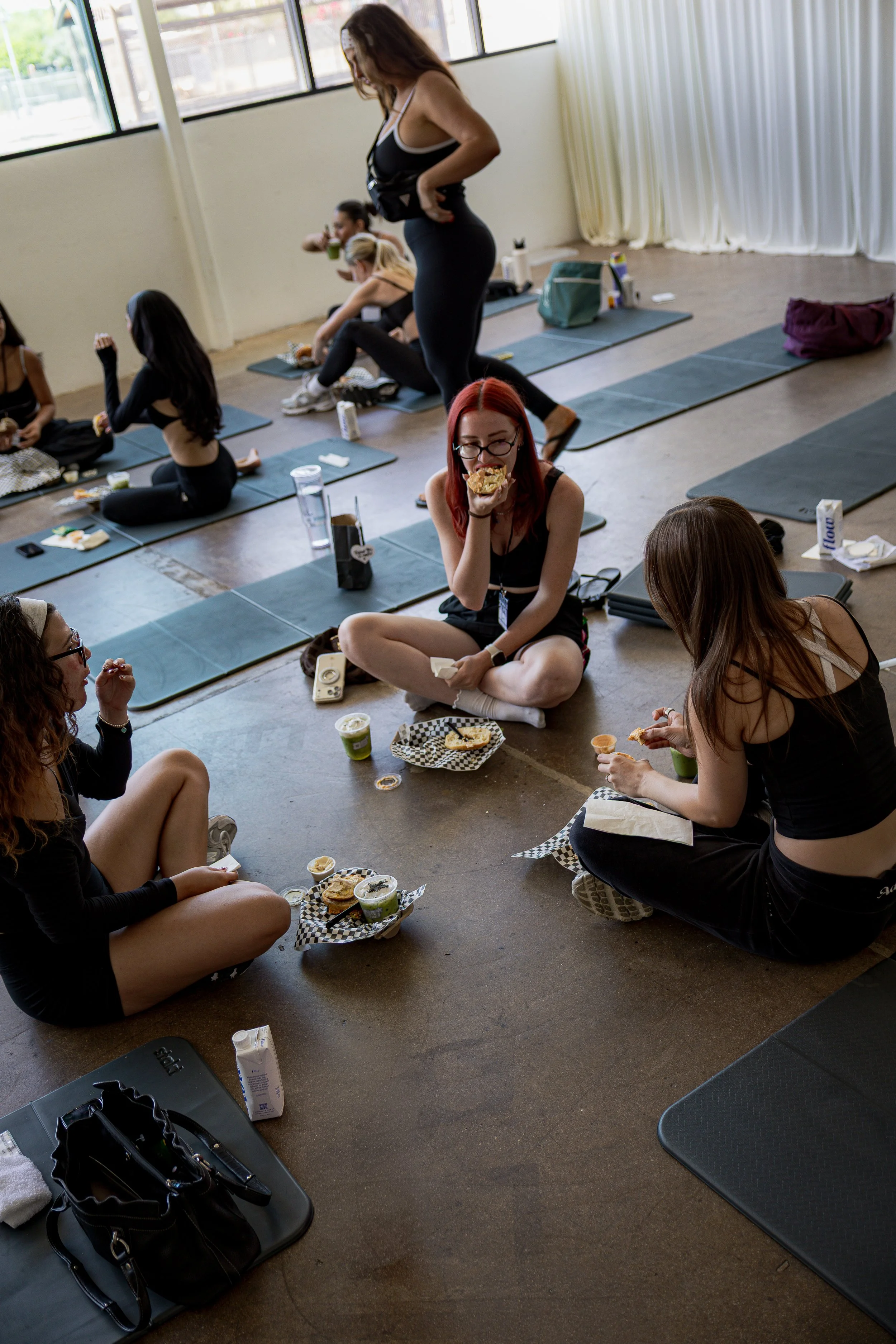 Women sitting on the floor with yoga mats, eating snacks, and socializing in a bright room with large windows.