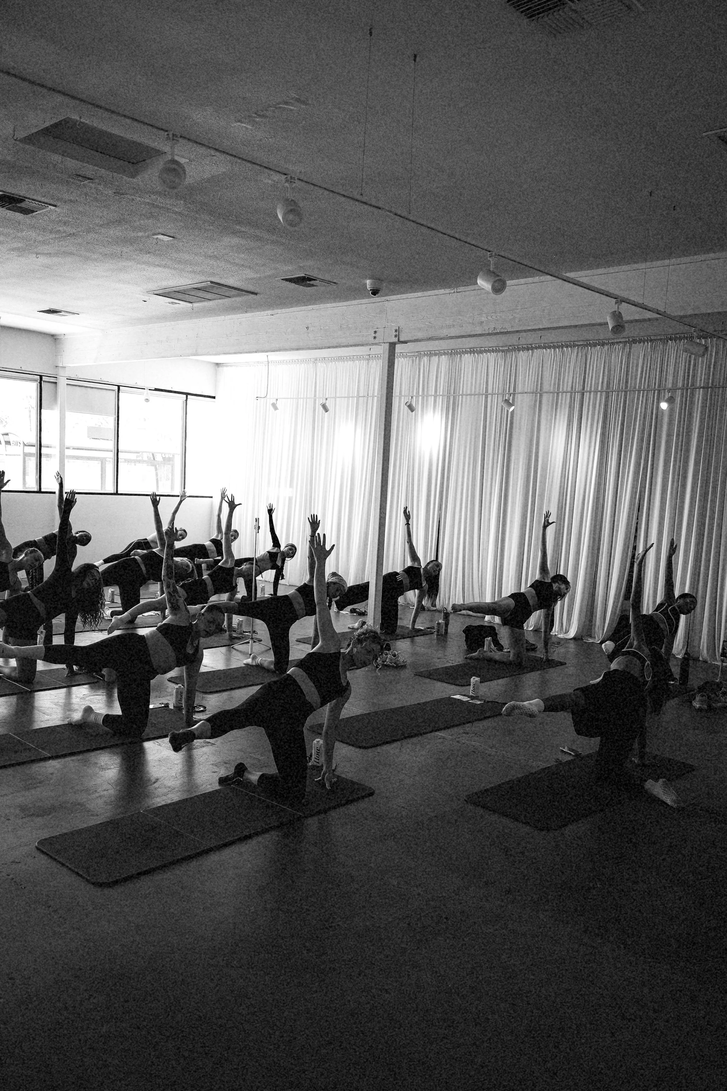 A large group of people practicing yoga in a studio with large windows and curtains, performing a pose with one arm extended upward and the opposite leg extended backward.