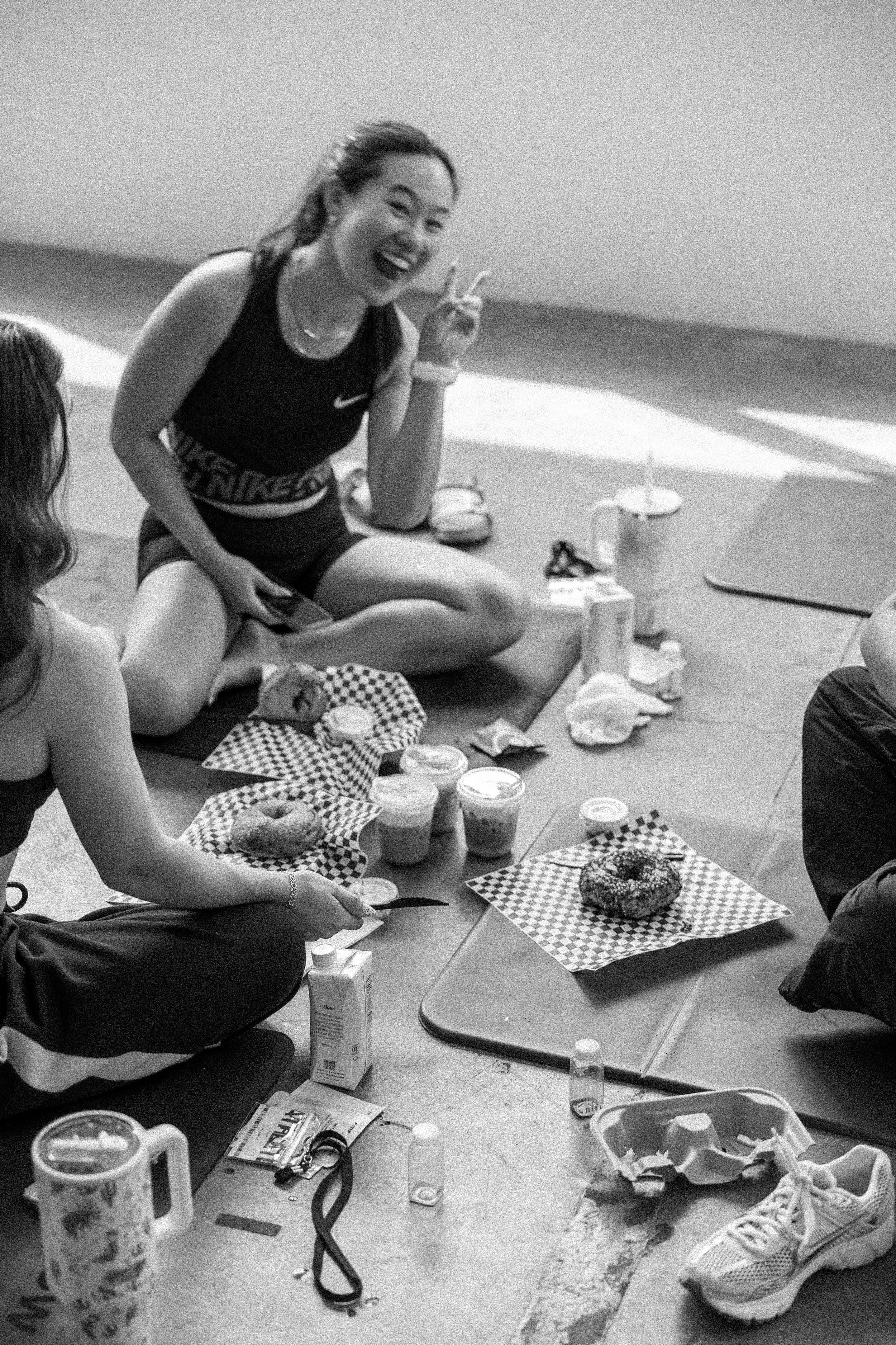 A woman in athletic clothing sitting on the floor, smiling, making a peace sign, with donuts and drinks on napkins in front of her, in an indoor setting.