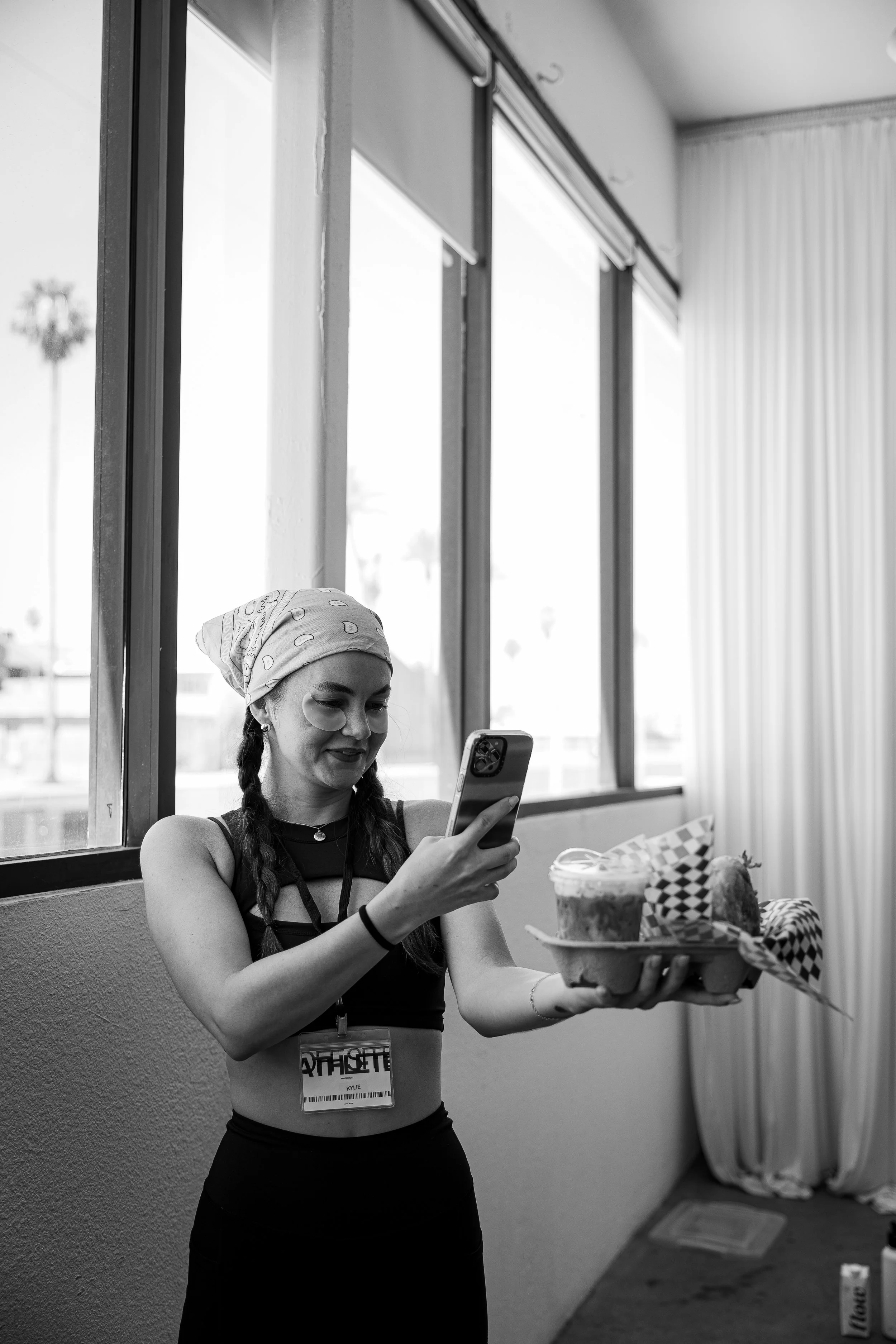 A young woman with braided hair, bandana, and a tank top is smiling and looking at her phone while holding a plate with a naked cake, a drink, and a wrapped gift, inside a room with large windows.